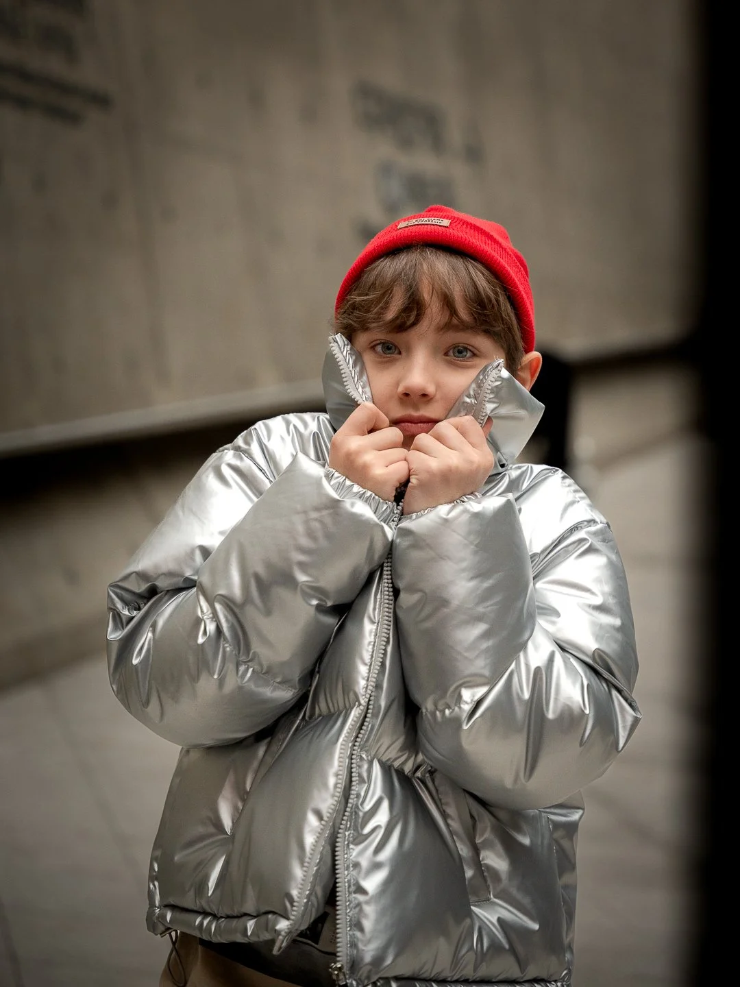Young boy wearing a red beanie and a shiny silver puffer jacket, standing indoors with a neutral expression.
