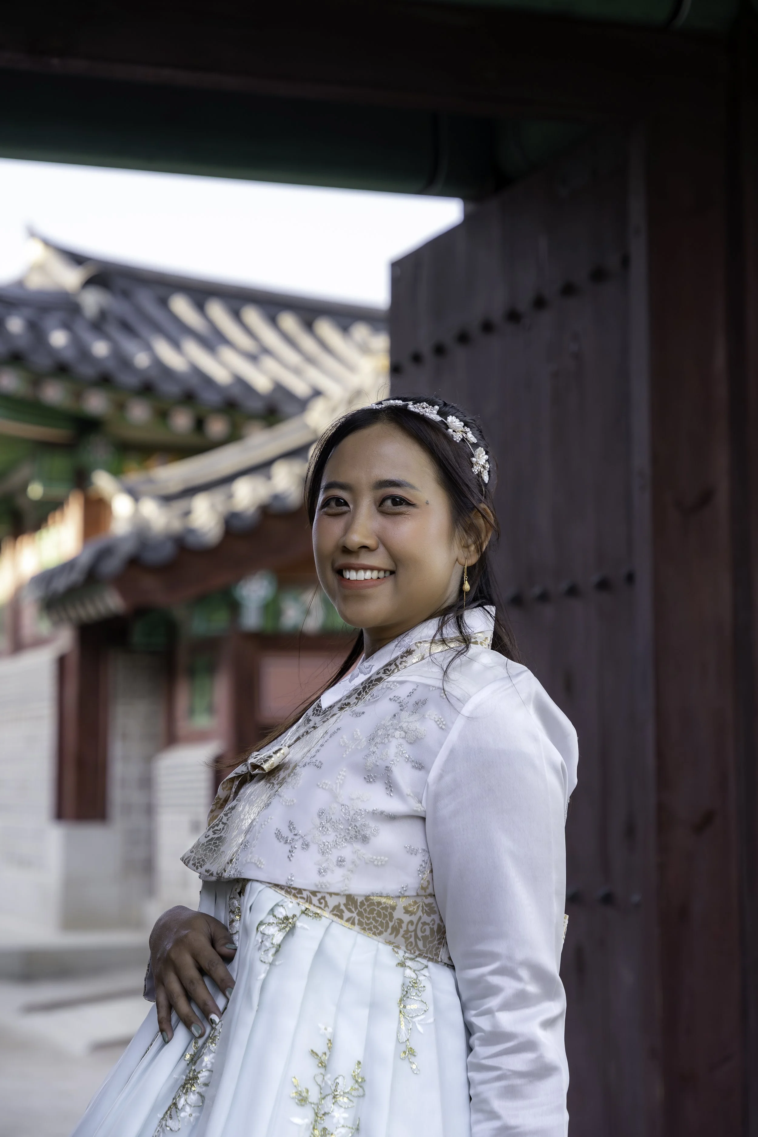 Young woman in traditional Korean wedding dress smiling at the camera outside a historic building.