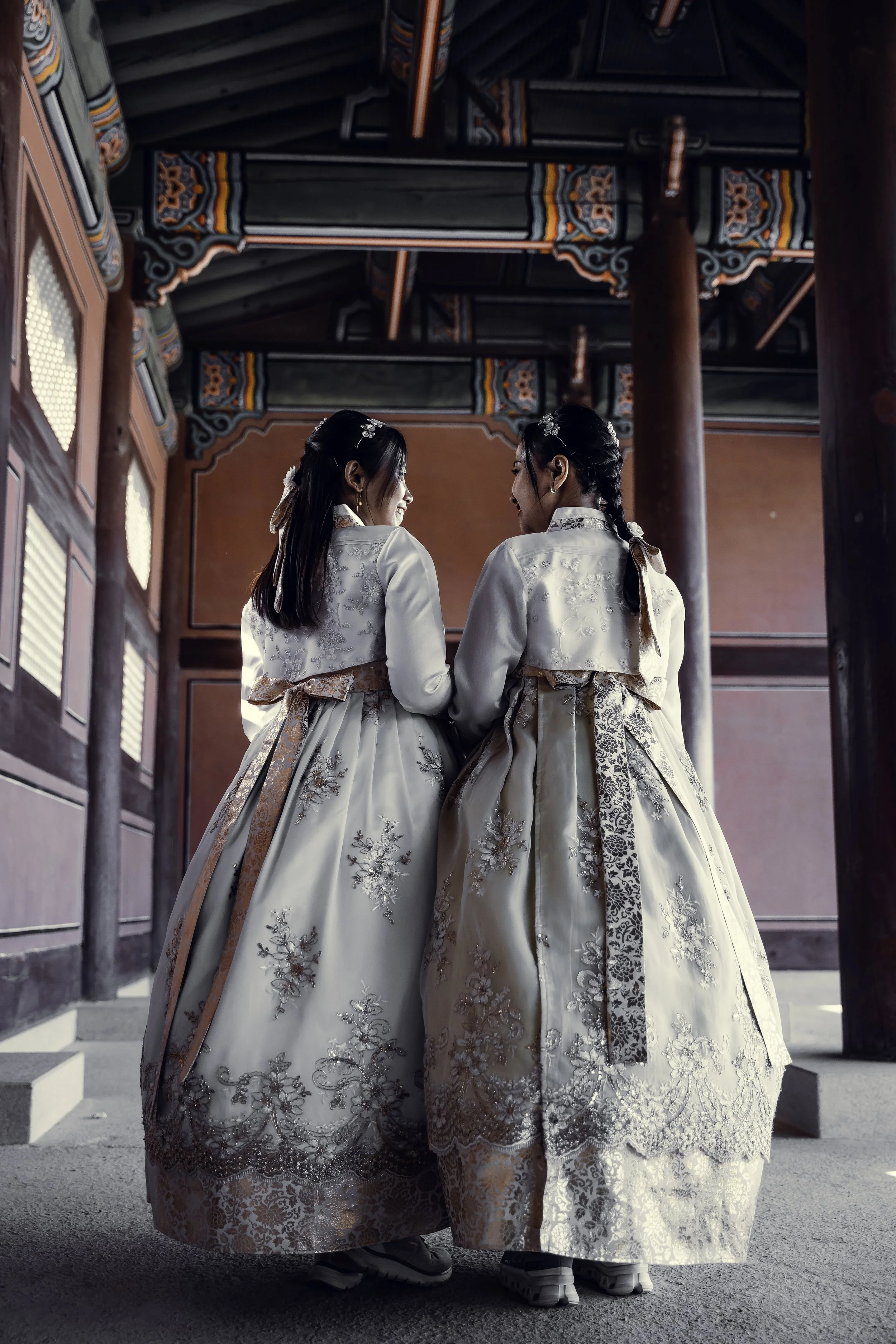 Two women dressed in traditional Korean hanbok standing together inside a historic building with ornate decor, facing each other.