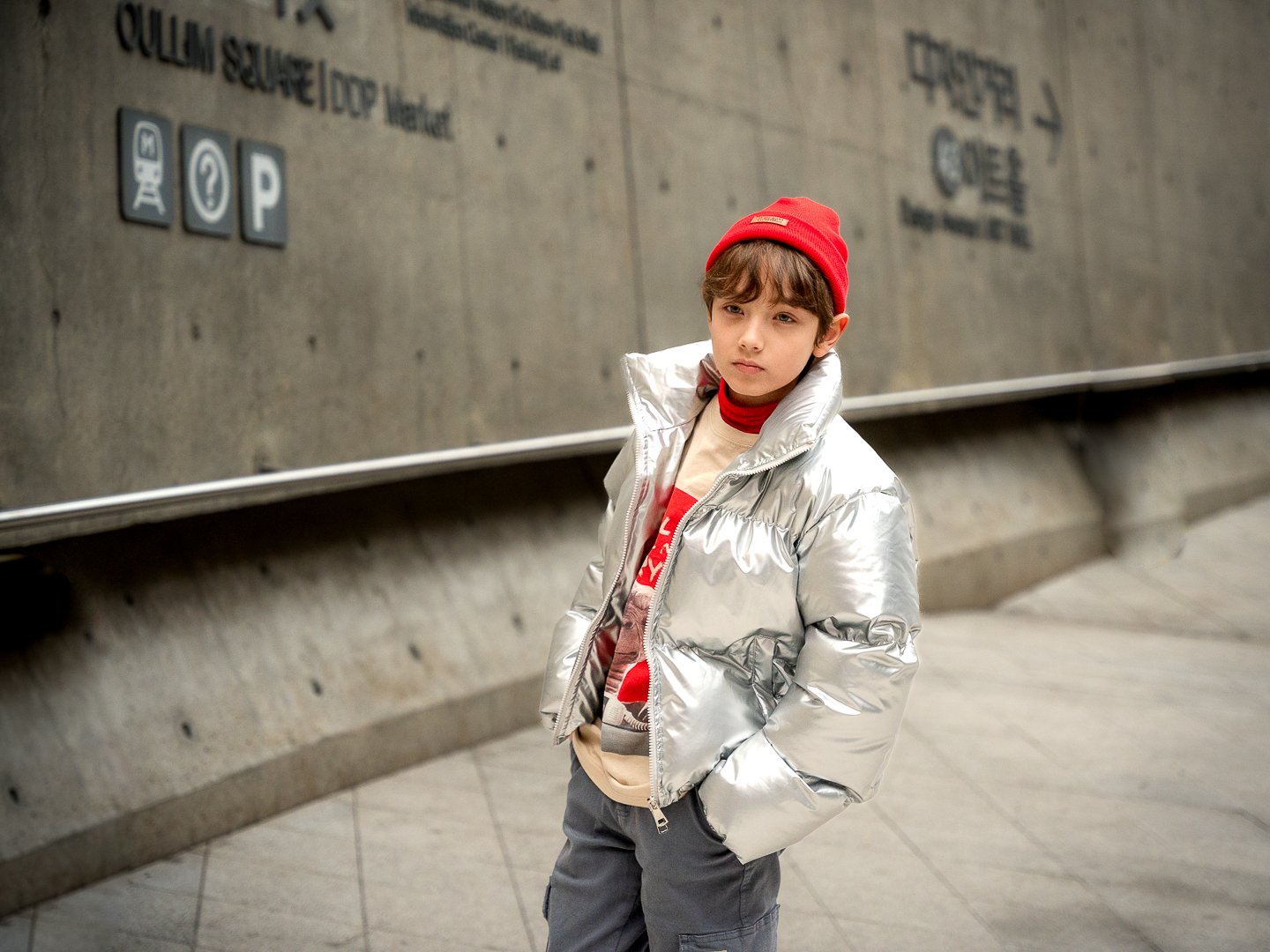 A young boy with brown hair, wearing a red beanie, a shiny silver puffer jacket, a beige graphic shirt, and gray pants, standing in an indoor urban setting near a concrete wall with signage.