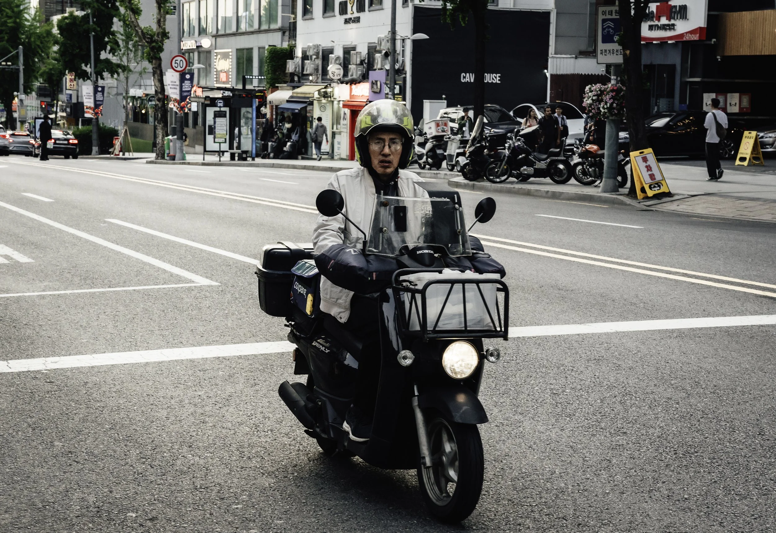 A man riding a black scooter on a city street, wearing a helmet and a white jacket, with stores and parked motorcycles in the background.