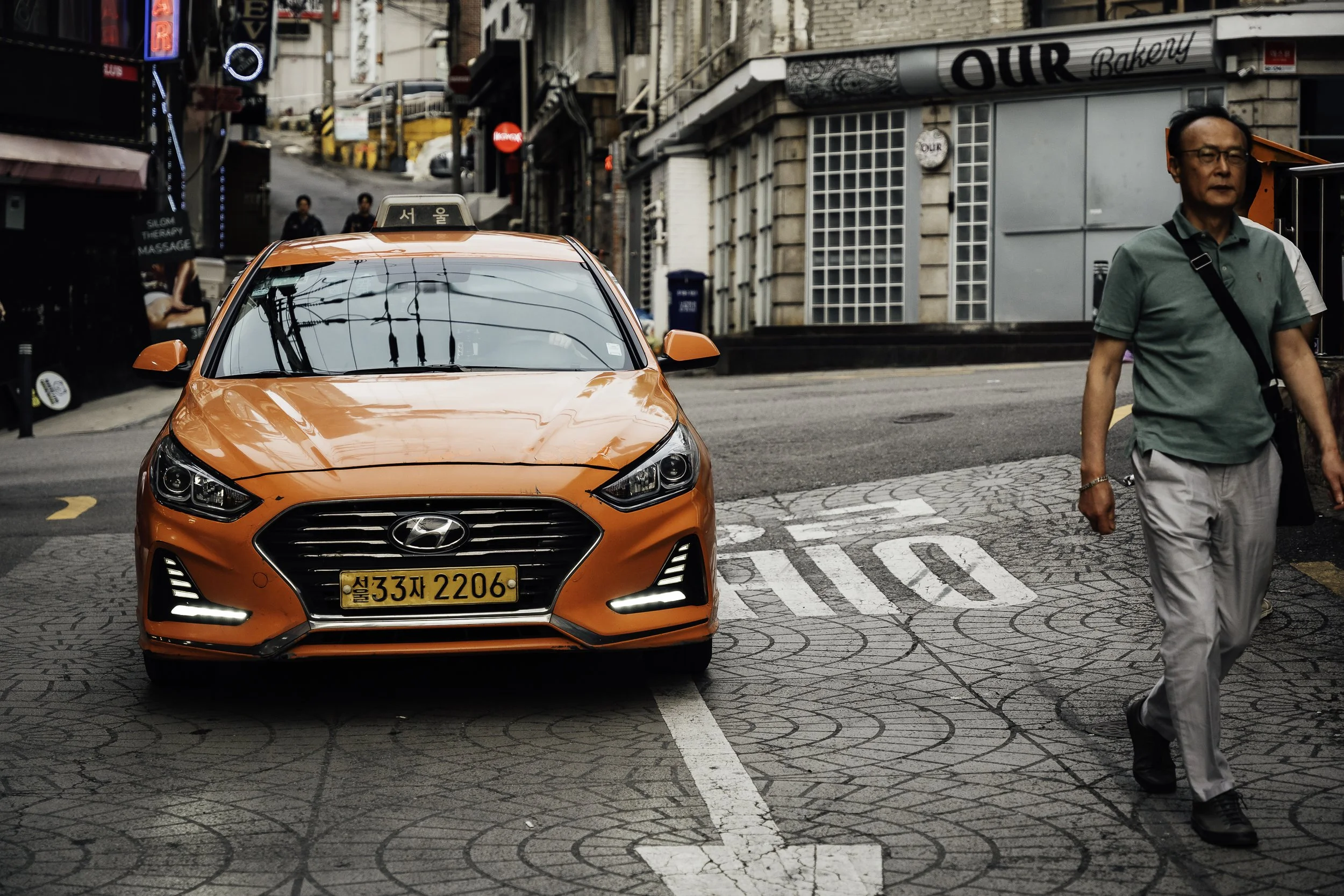 An orange Hyundai taxi with Korean license plate parked on a city street crosswalk, with a man in a green shirt and light-colored pants walking past.