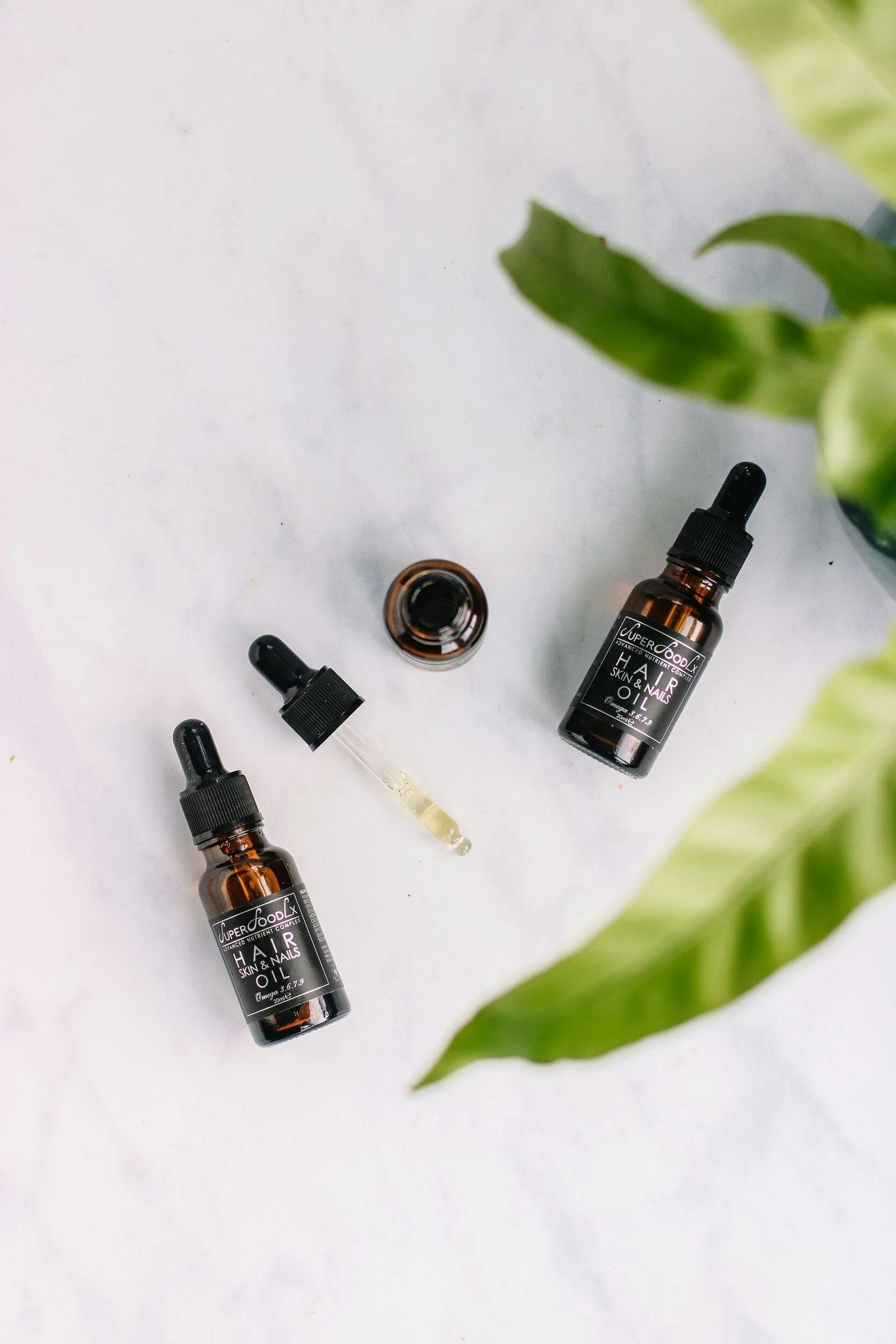 Two small bottles of skin and nail oil with dropper caps, one open with a dropper filled with oil, placed on a white surface surrounded by green leaves.