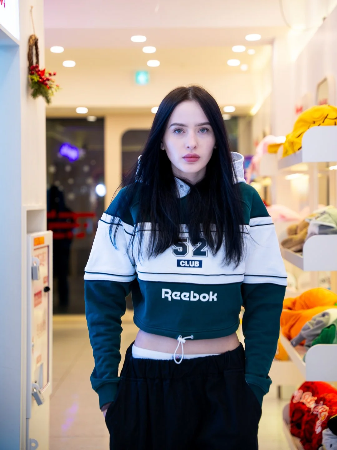 A young woman with long black hair and fair skin standing inside a store, wearing a cropped Reebok hoodie with the number 52 and a club logo, black pants, and with her hands in her pockets.