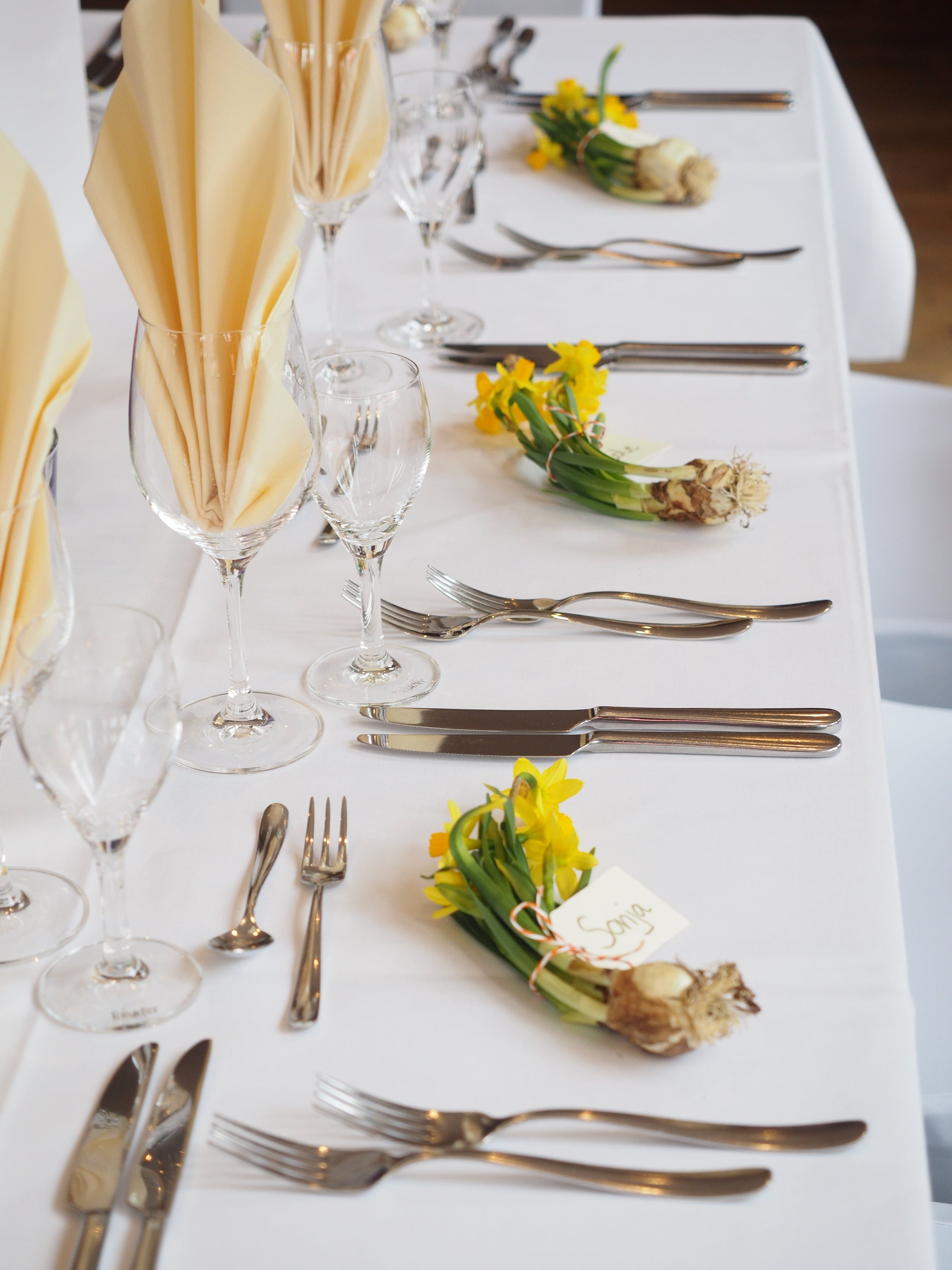 Elegant dining table with yellow flowers, folded napkins in wine glasses, and neatly arranged silverware on a white tablecloth.