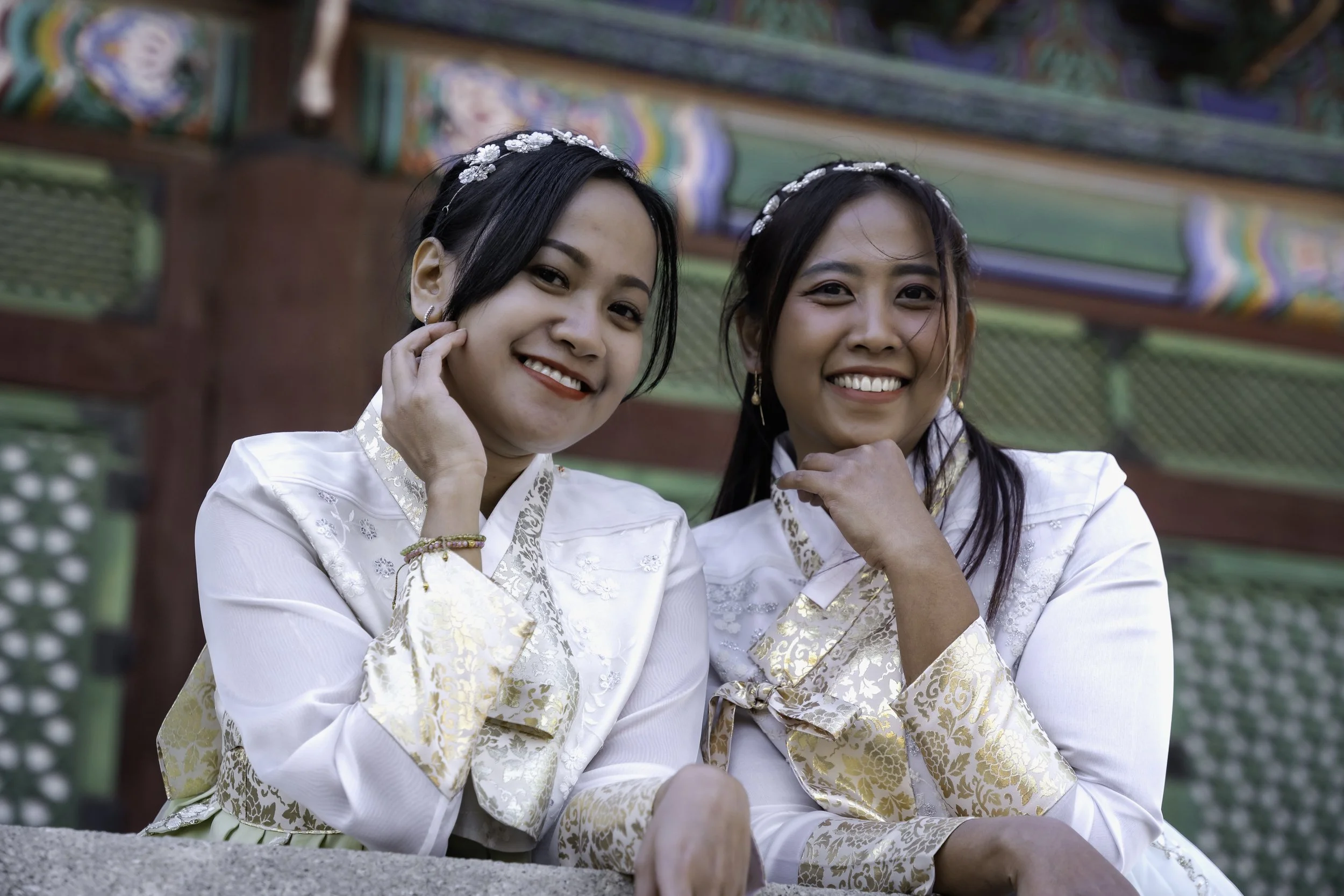 Two young women wearing traditional white and gold dresses with floral headbands, smiling and posing in front of a colorful temple.