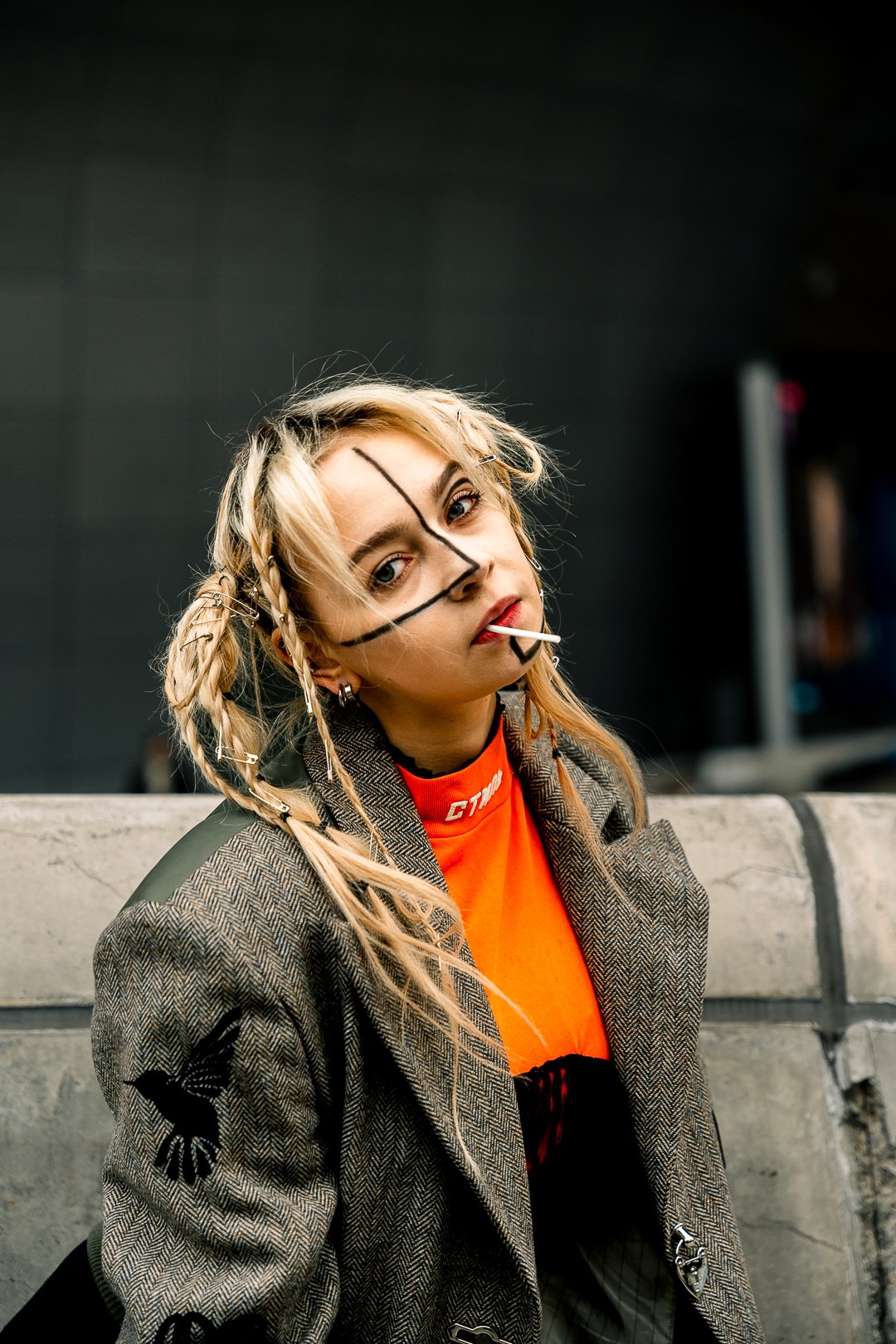 A young woman with blonde hair styled in braids and wearing a blazer with a dove patch, an orange shirt, and dark makeup, leaning against a concrete surface in an urban setting, with a lollipop in her mouth and face paint in black lines.