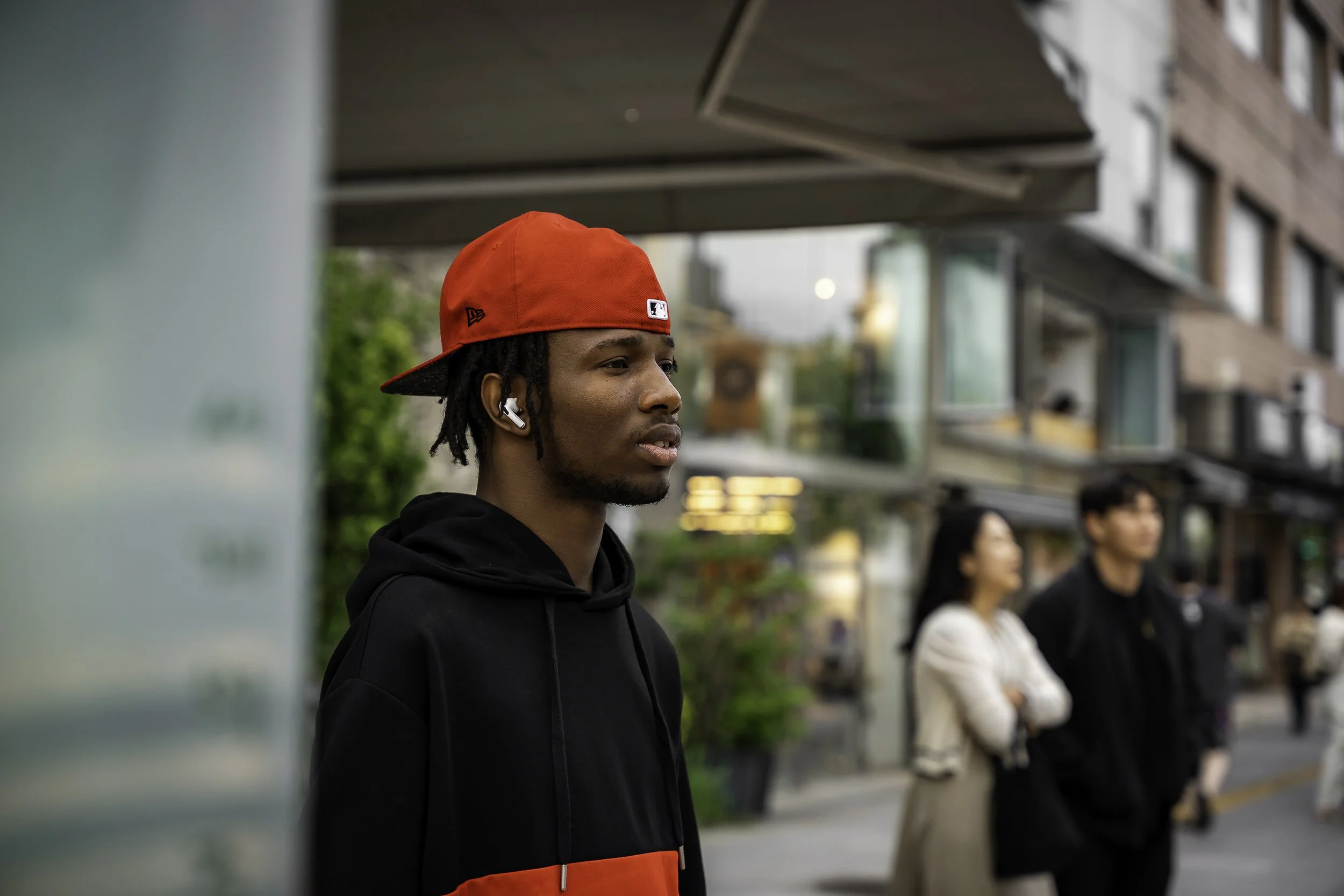 Young man with dreadlocks wearing a red baseball cap, black hoodie, and wireless earbuds, standing on a city sidewalk.
