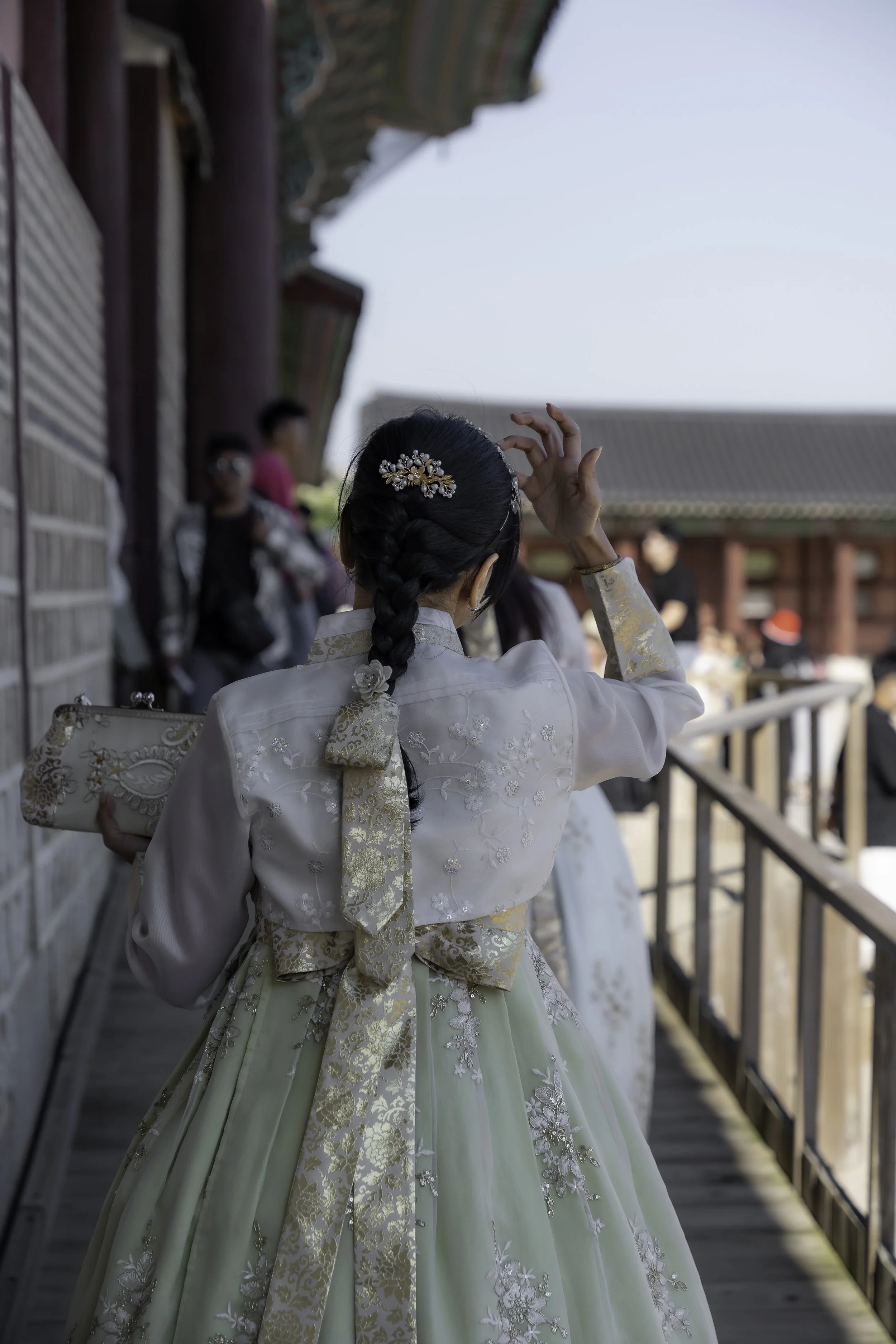 A woman dressed in traditional Korean hanbok walking on a wooden pathway with a group of people in the background at a historical site.