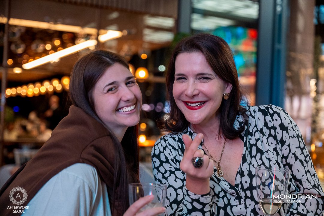 Two women smiling and making a heart gesture with their hands at a social event, holding glasses of wine.