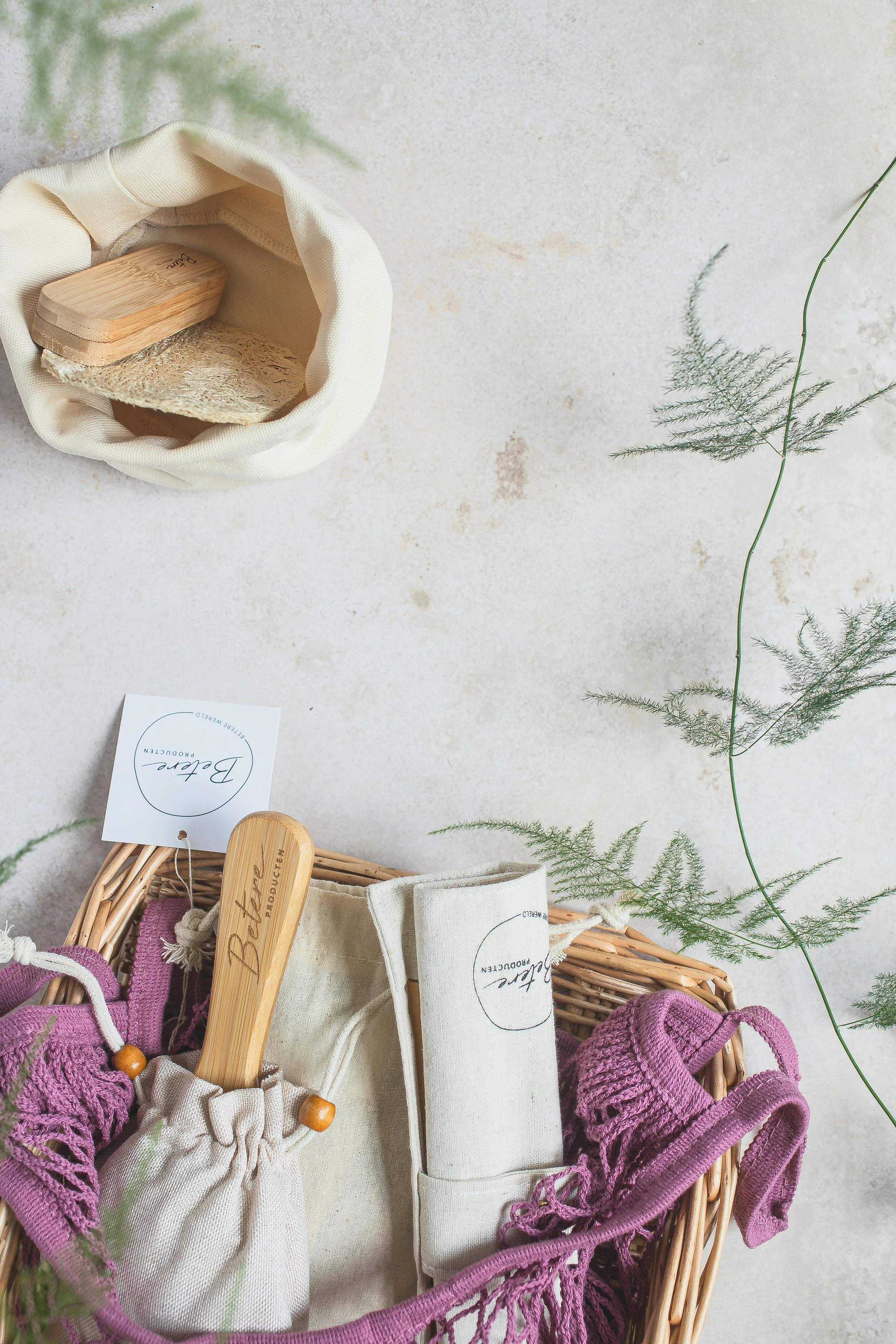 A collection of eco-friendly self-care items including a wooden brush, a fabric pouch, and cloth slippers in a woven basket, with a small bag and tags, placed on a neutral textured surface with green foliage.