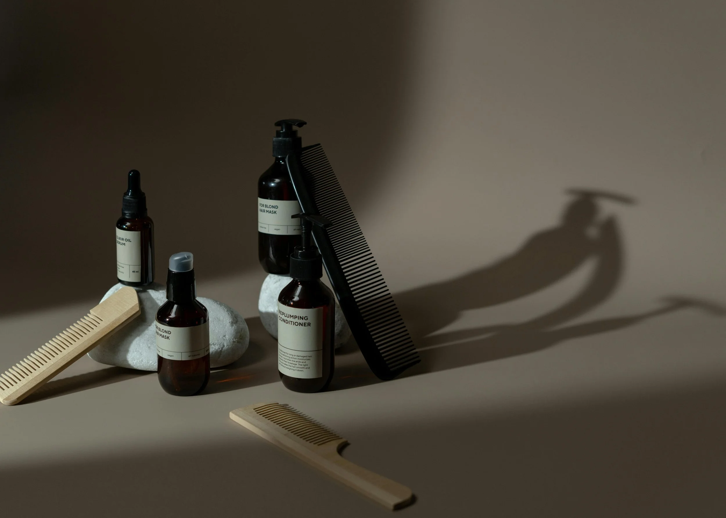 Hair care products including bottles of hair mask, conditioner, hair oil, a black hair comb, and wooden hairbrushes arranged on rocks with shadows cast on a beige background.