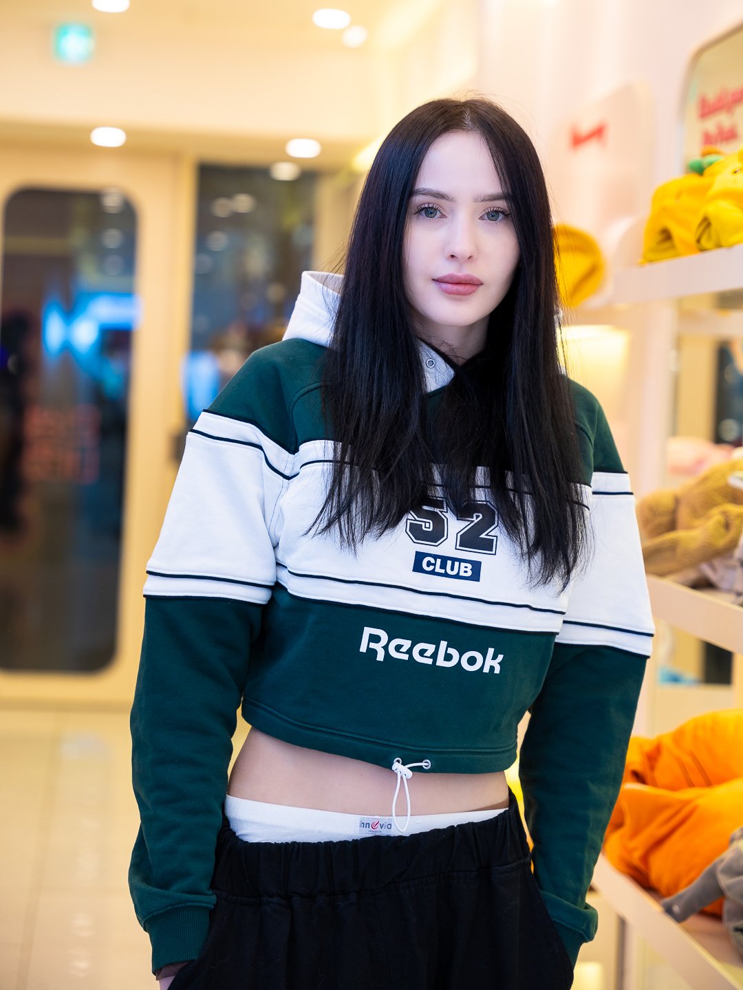 A young woman with long dark hair and light skin standing inside a store, wearing a green and white cropped hoodie with Reebok branding, black pants, and a white waistband visible.