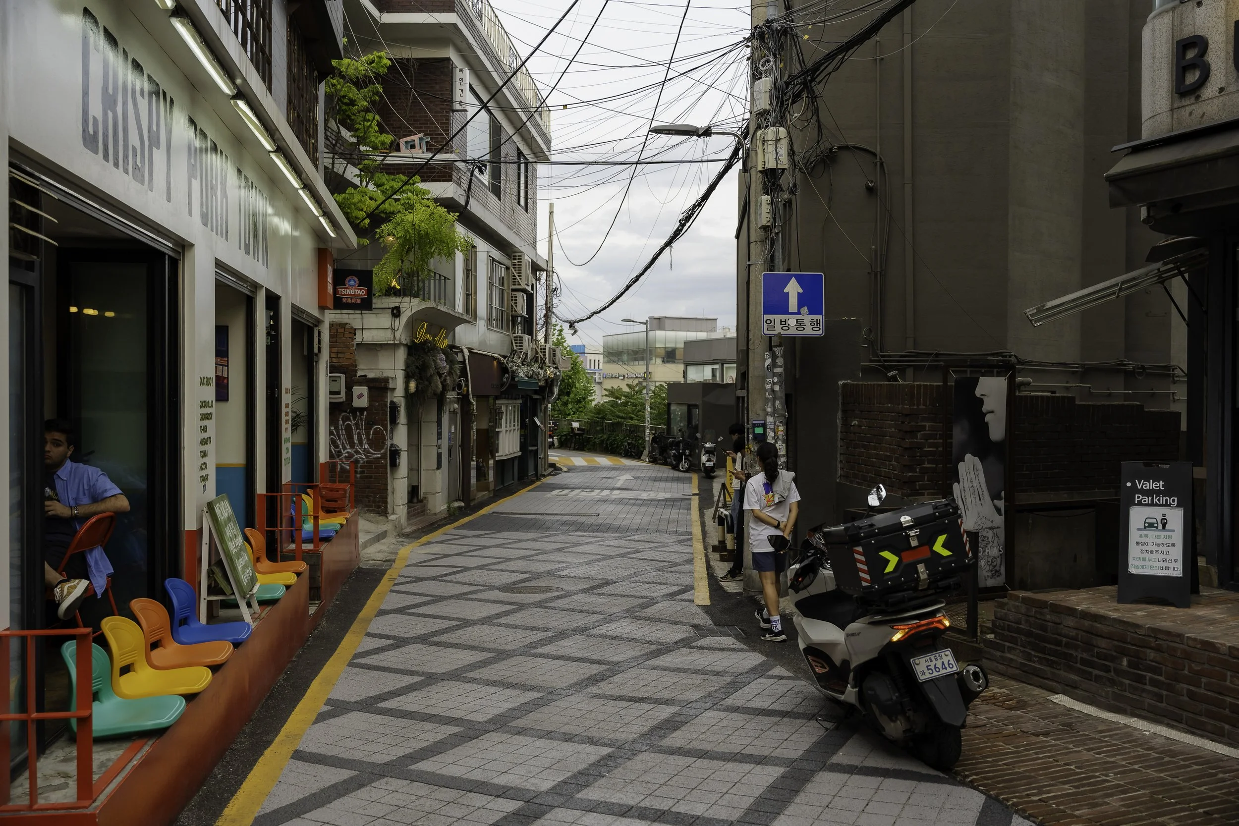 A narrow urban street with colorful chairs outside a cafe on the left, and pedestrians on the right sidewalk, with a parked scooter and various signage, including Korean characters and a posted valet parking sign.