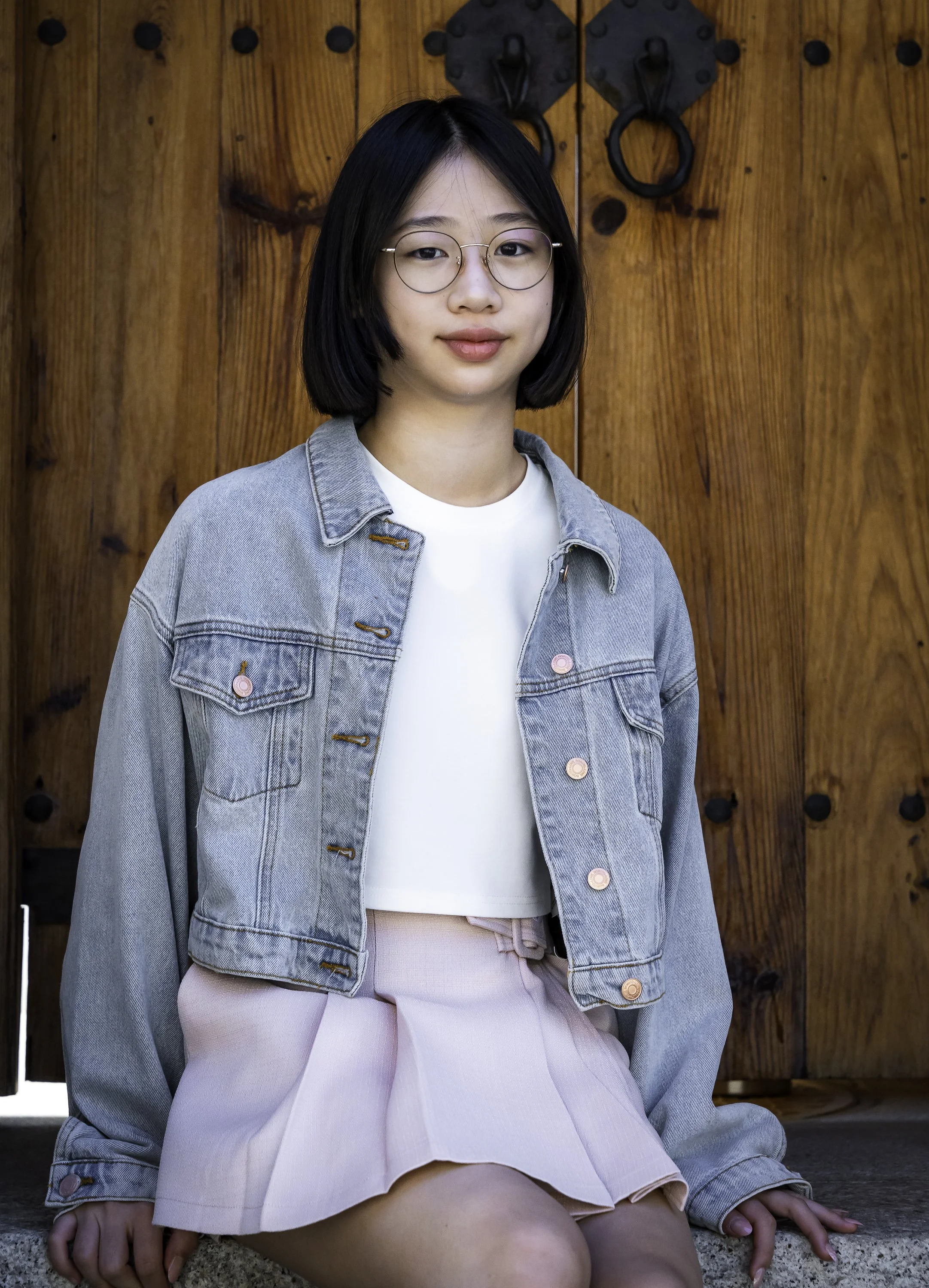 A young woman with short black hair and glasses sitting in front of a wooden door with black iron details, wearing a light denim jacket, white crop top, and pink pleated skirt.