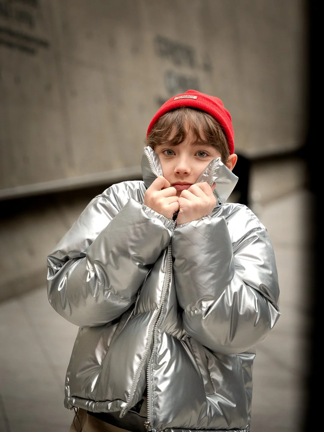 Young boy wearing a red beanie and shiny silver puffer jacket, standing outdoors against a blurred background.