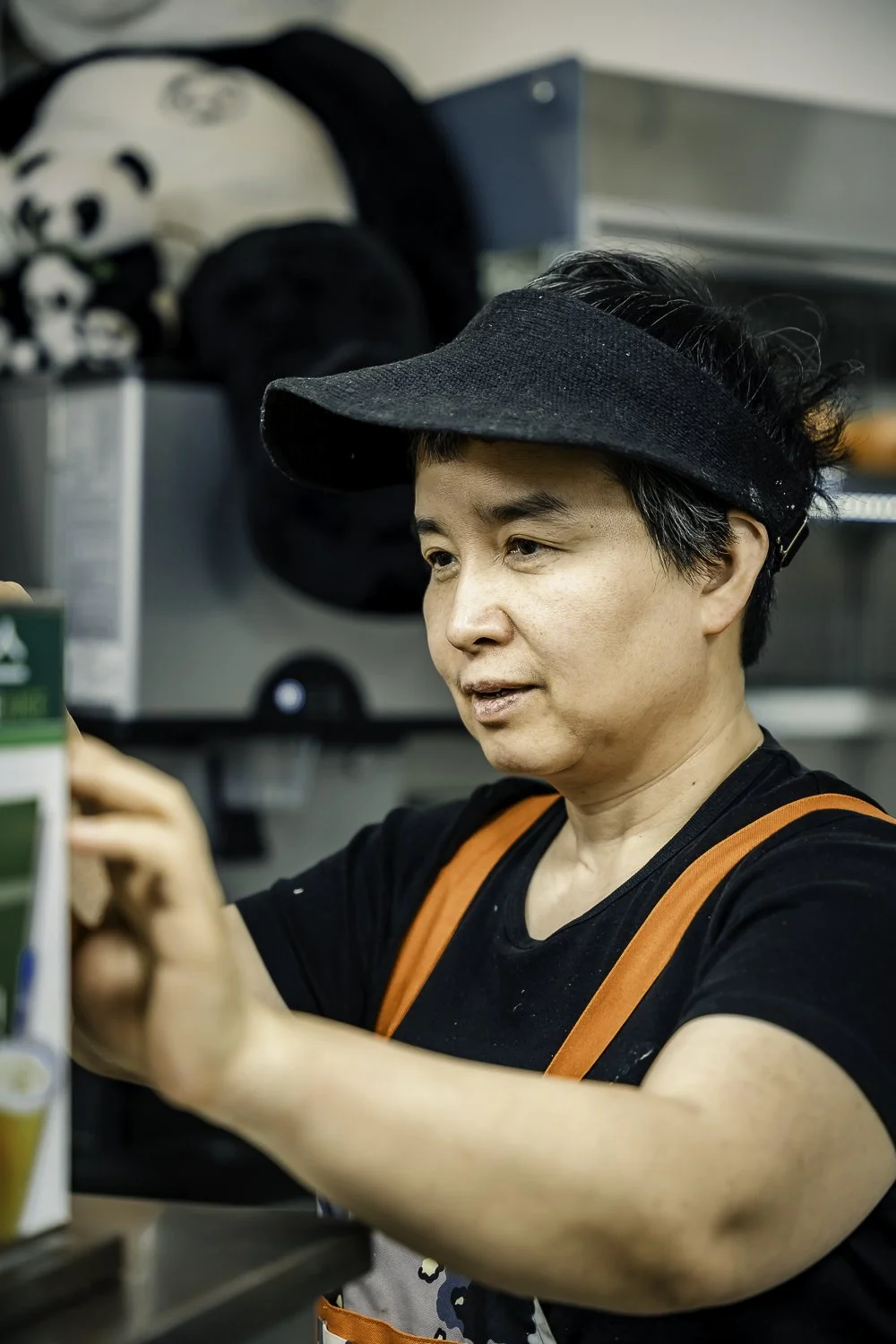 A woman in a black visor and black shirt with orange suspenders taking a selfie in a kitchen or cafe setting.