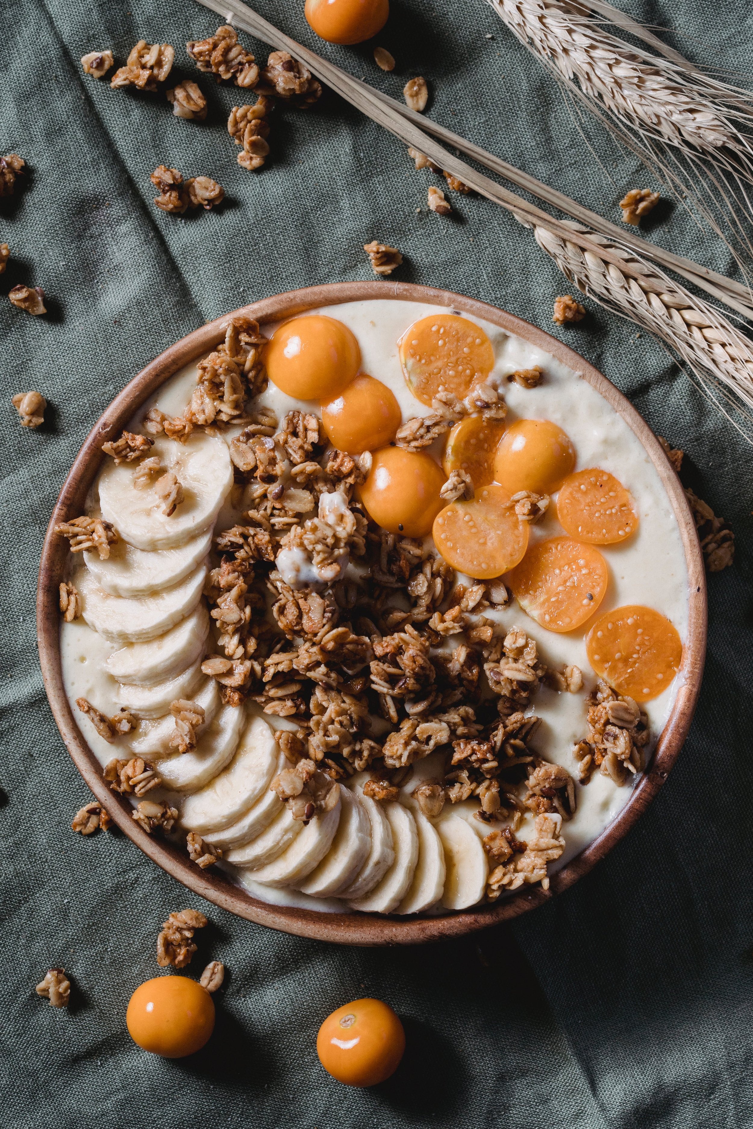 A bowl of yogurt topped with sliced bananas, granola, and yellow cherry tomatoes on a dark cloth with wheat stalks.