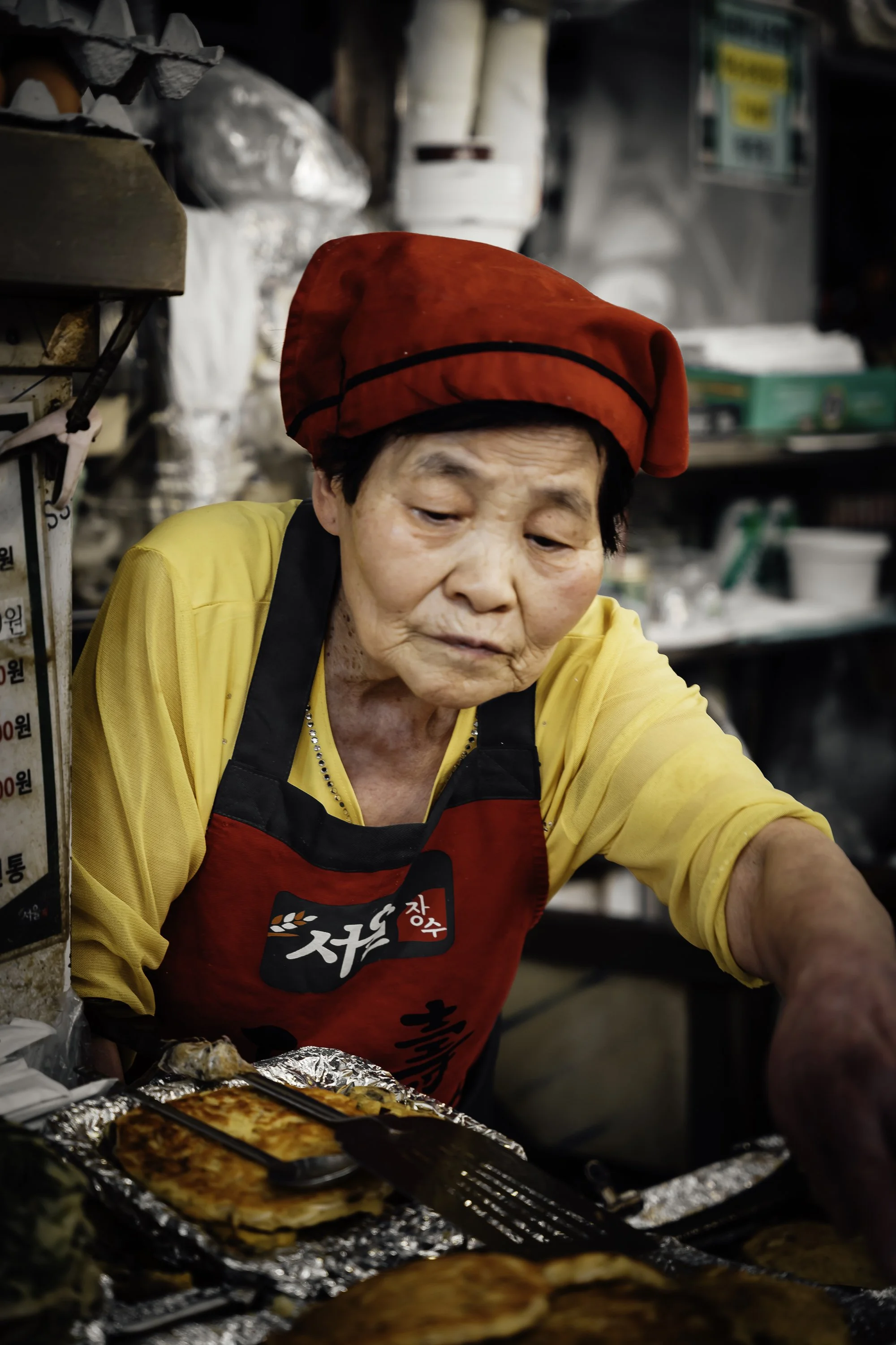 An elderly woman wearing a red hat, yellow shirt, and black-and-red apron preparing food at a street stall with grilled items in foil containers.