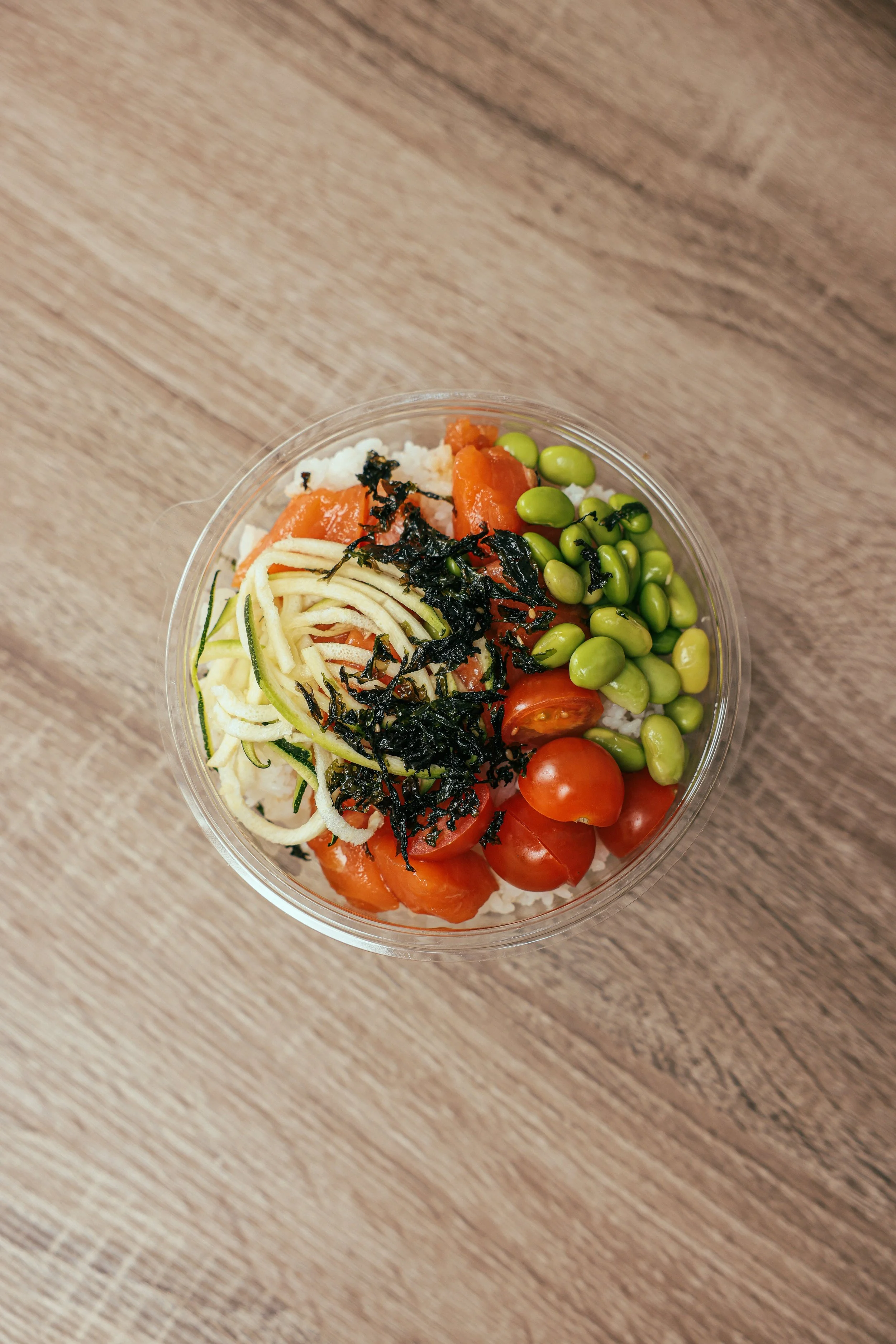 A clear bowl of salad with cherry tomatoes, edamame beans, shredded cucumber, shredded cheese, shredded seaweed, and rice, on a wooden surface.