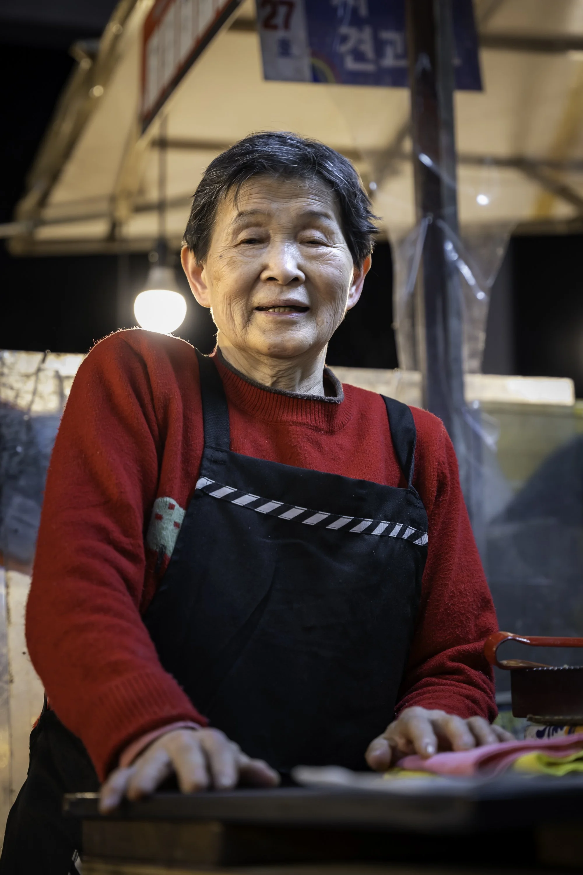 Older woman with short black hair wearing a red sweater and black apron, standing behind a table at an outdoor market or food stall.