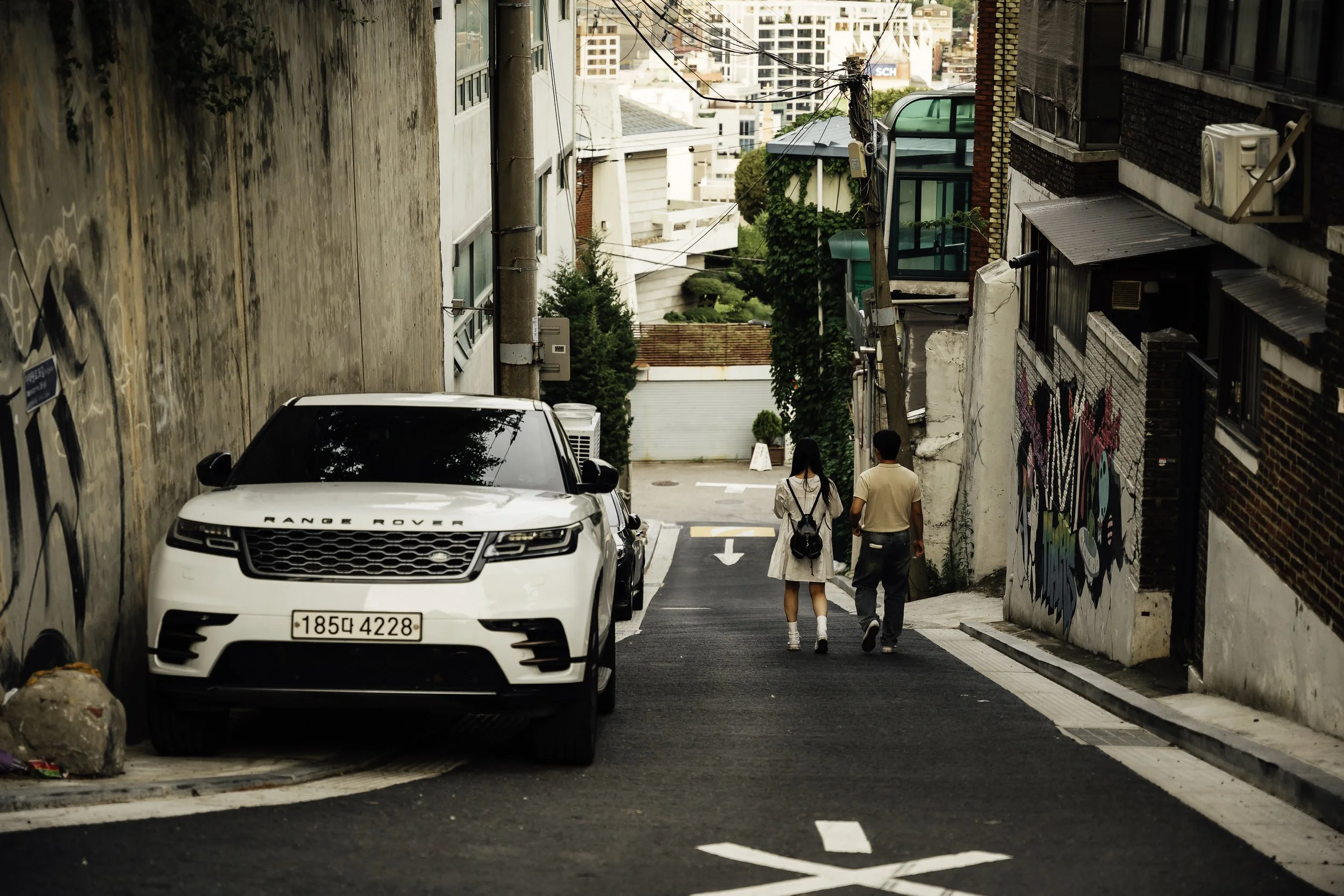 Two people, a woman and a man, walk down a narrow street lined with parked cars, graffiti, and buildings in an urban area.