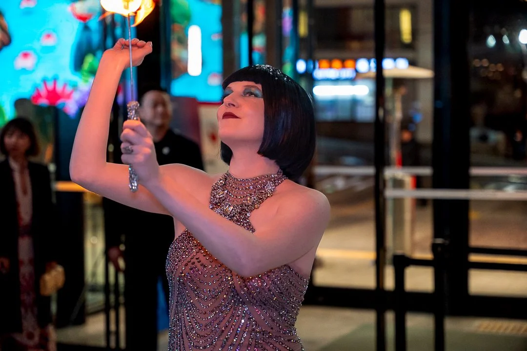 A woman with a short black bob haircut and glamorous makeup wearing a strapless, beaded evening gown and multiple necklaces, holding a sparkler inside at night with bright city lights and people in the background.