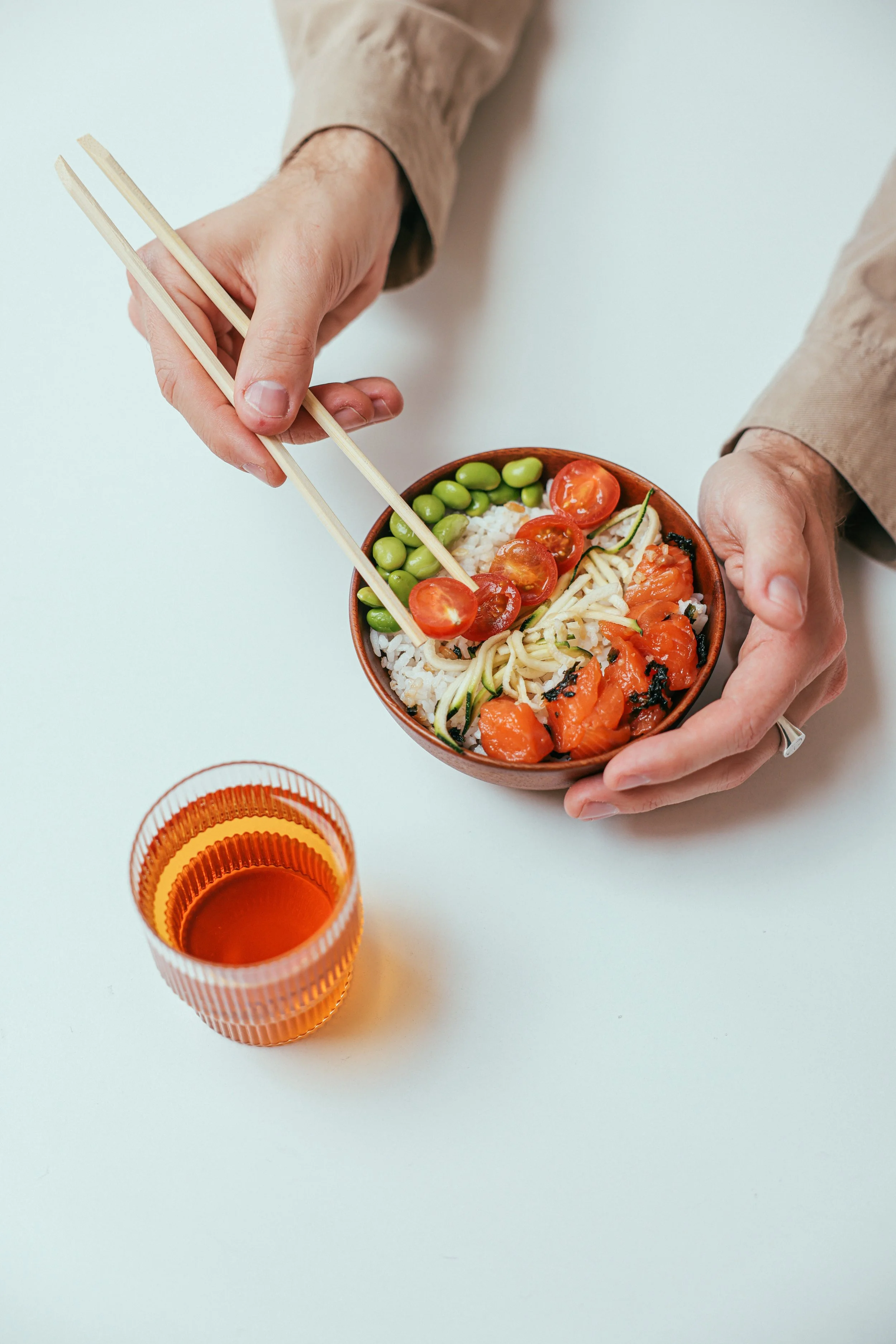 Person holding a bowl of sushi with chopsticks, a glass of tea on white table.