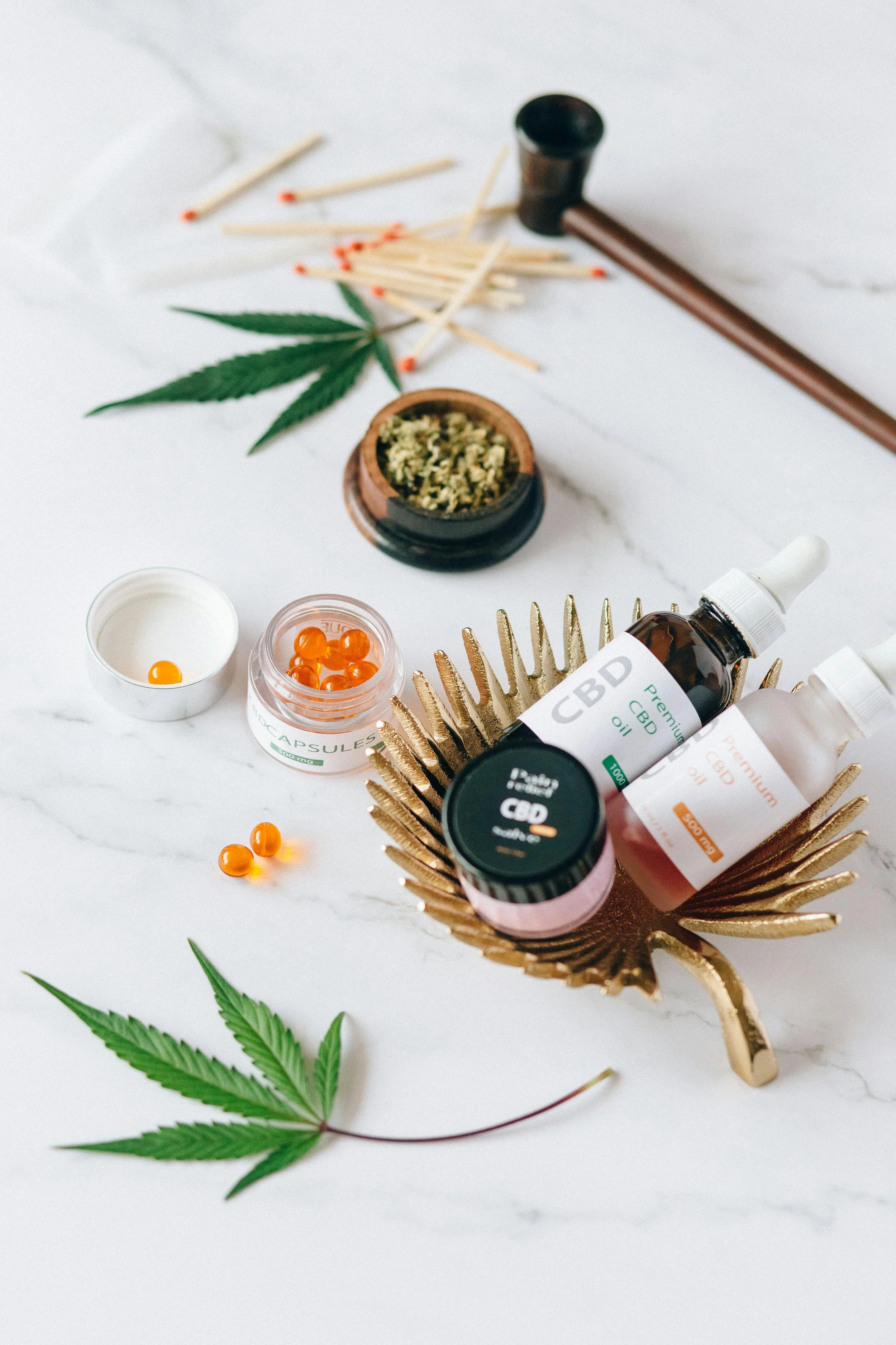 CBD products, including oil bottles, capsules, and creams, along with cannabis leaves, matches, and dried herbs, displayed on a marble surface.