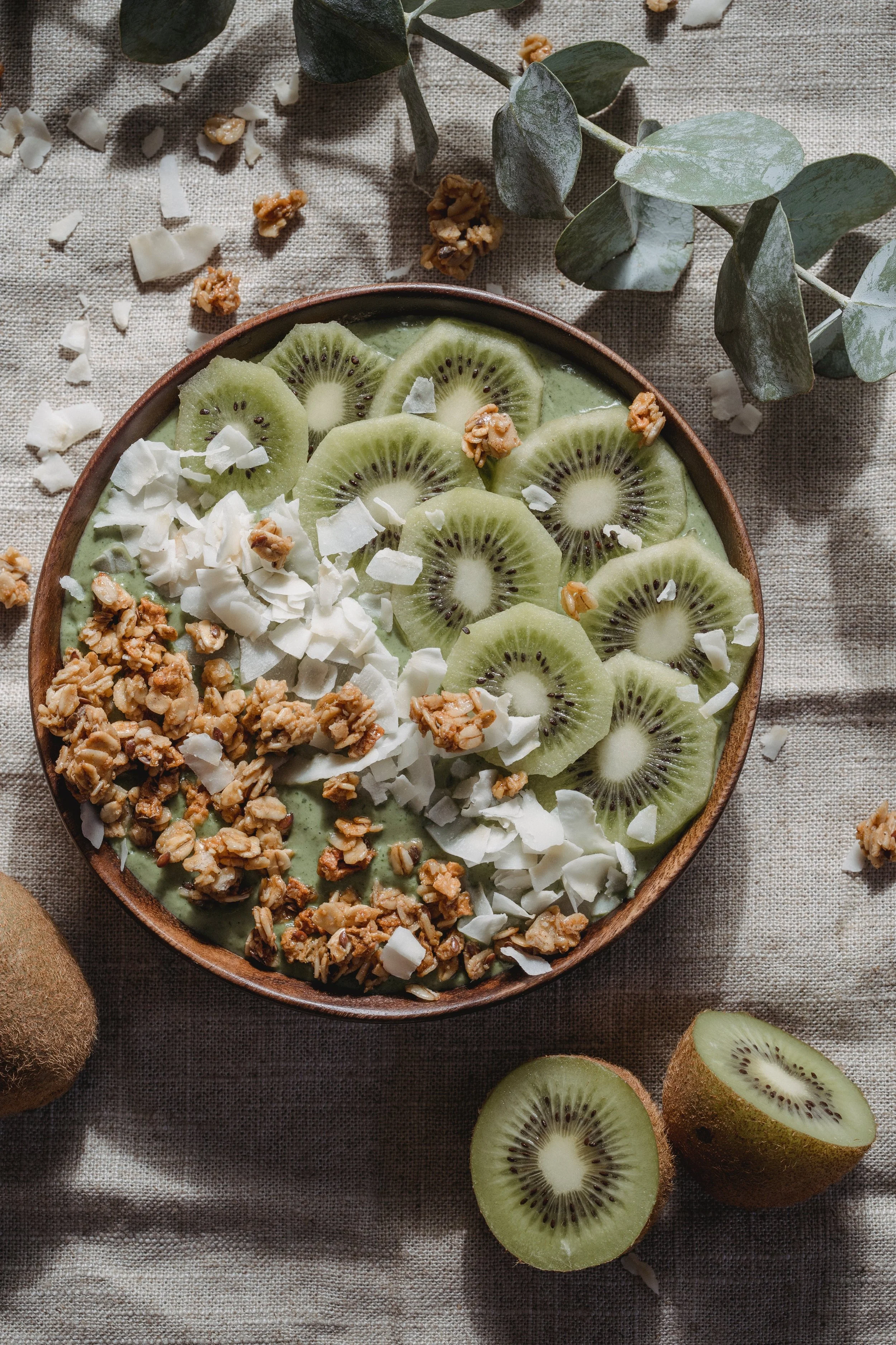 A bowl of green smoothie topped with sliced kiwi, granola, and white coconut flakes, placed on a neutral cloth with extra kiwi and some green leaves around it.