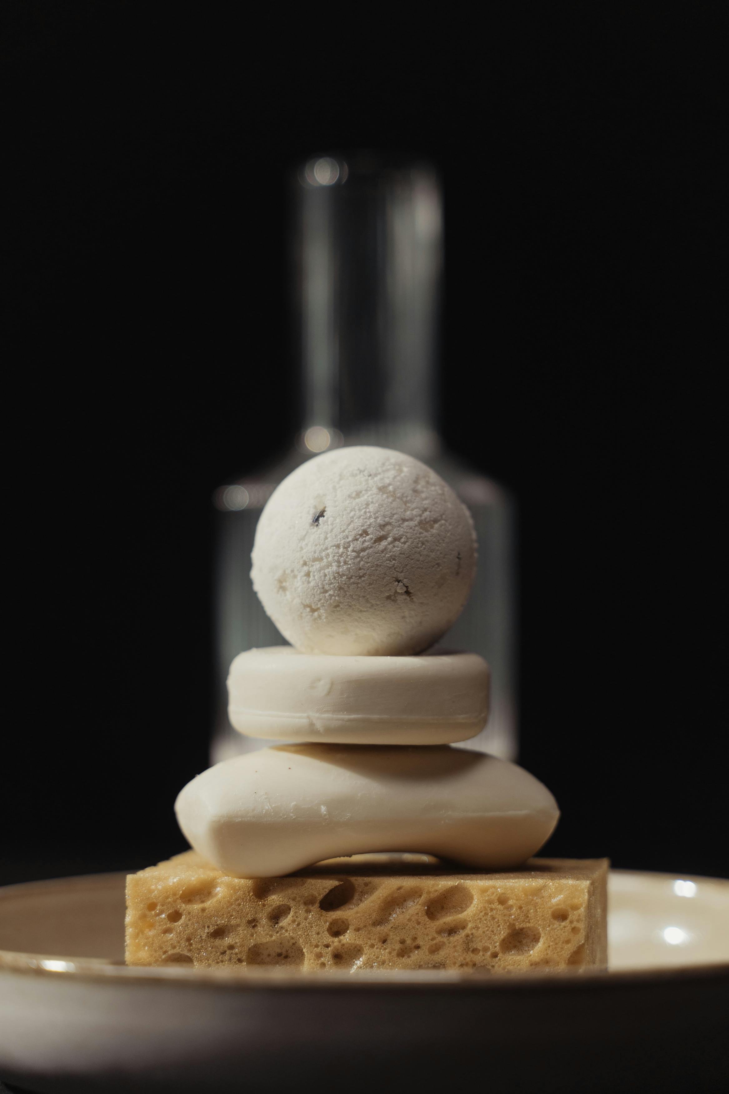 A stack of white objects, including a sponge, a round soap, a bar of soap, and a spherical bath bomb, arranged on a plate with a dark background.