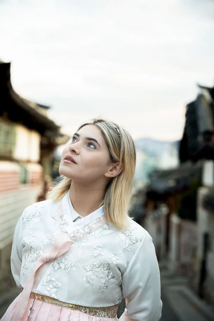 Young woman with blonde hair and a light pink and white dress standing on a street with traditional Asian buildings behind her.
