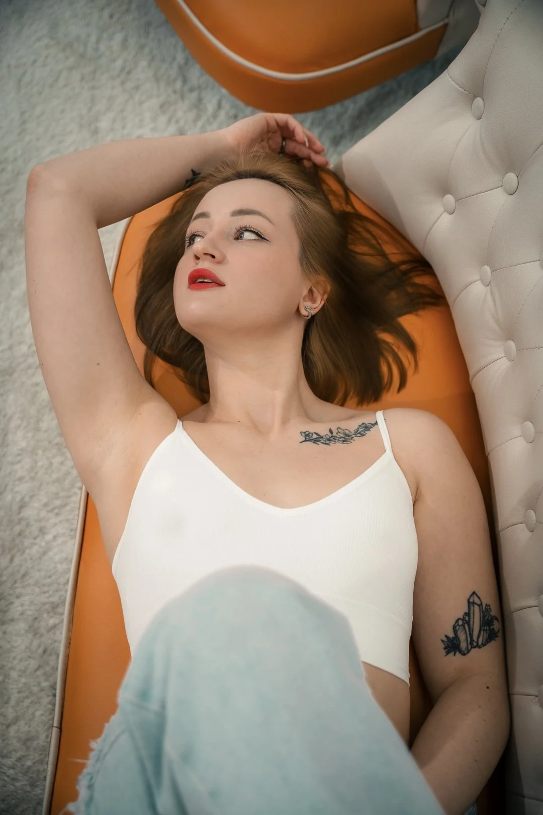 A young woman with fair skin, red hair, and tattoos, lying on an orange and white couch, wearing a white tank top, looking up with her left hand behind her head.
