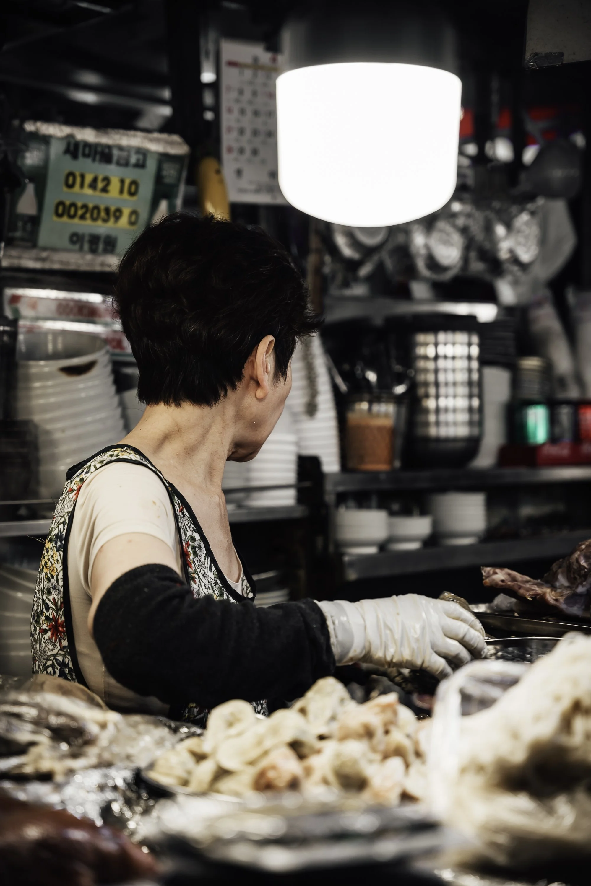 A woman wearing gloves preparing meat at a market stand with plates and containers in the background.