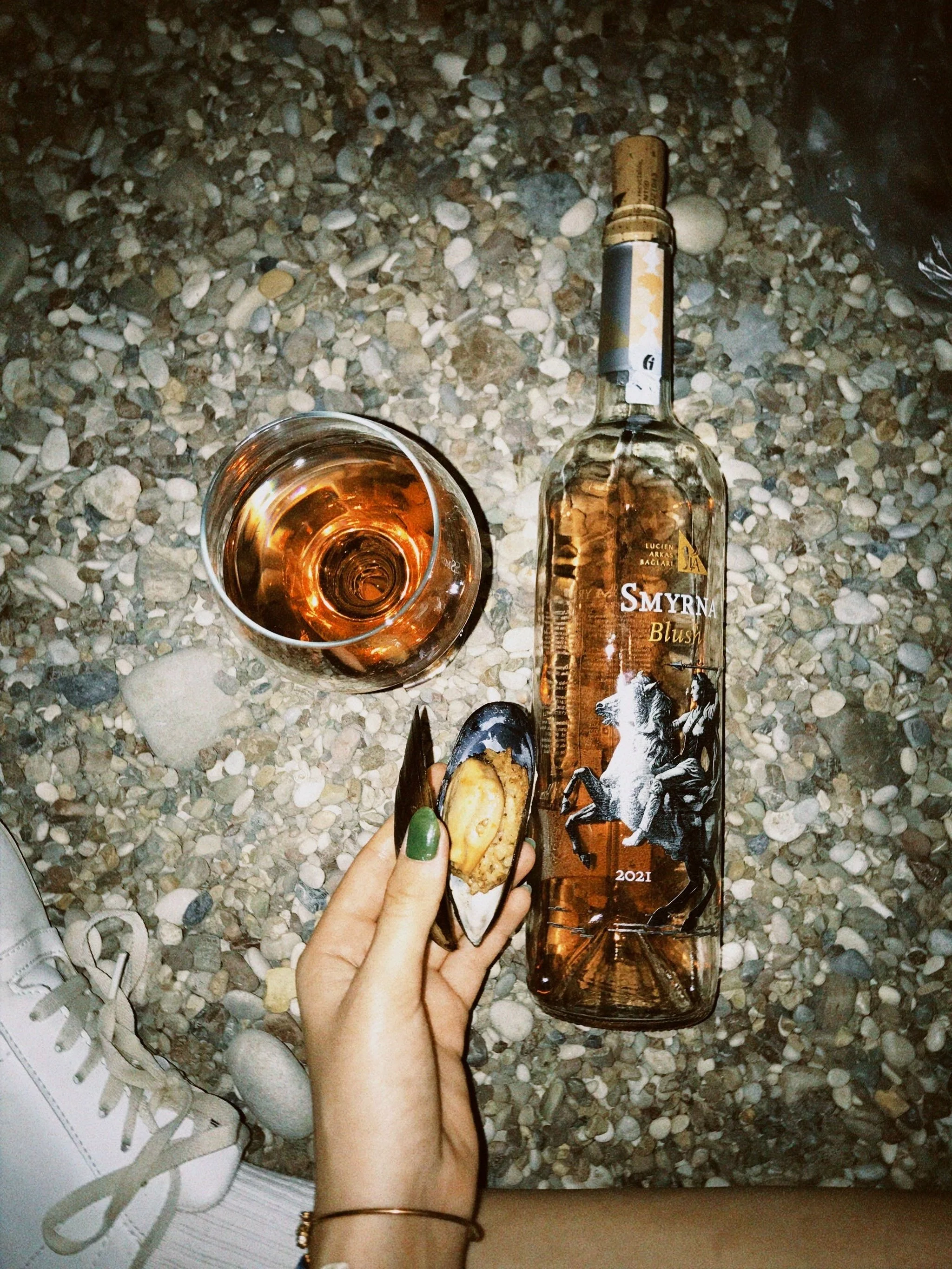 A hand with green-painted nails holding a shell with a piece of bread, next to a glass of rosé wine and a bottle of Smyrna Blush rosé wine with artistic label, on a pebbled ground.
