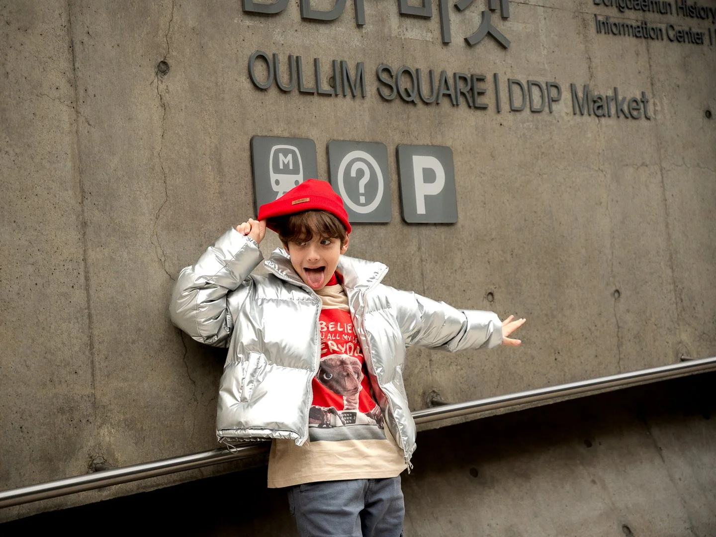 A young boy in a shiny silver puffer jacket and red beanie hat standing at a train station, making a funny face with his tongue out and arms outstretched, in front of a concrete wall with station sign symbols.