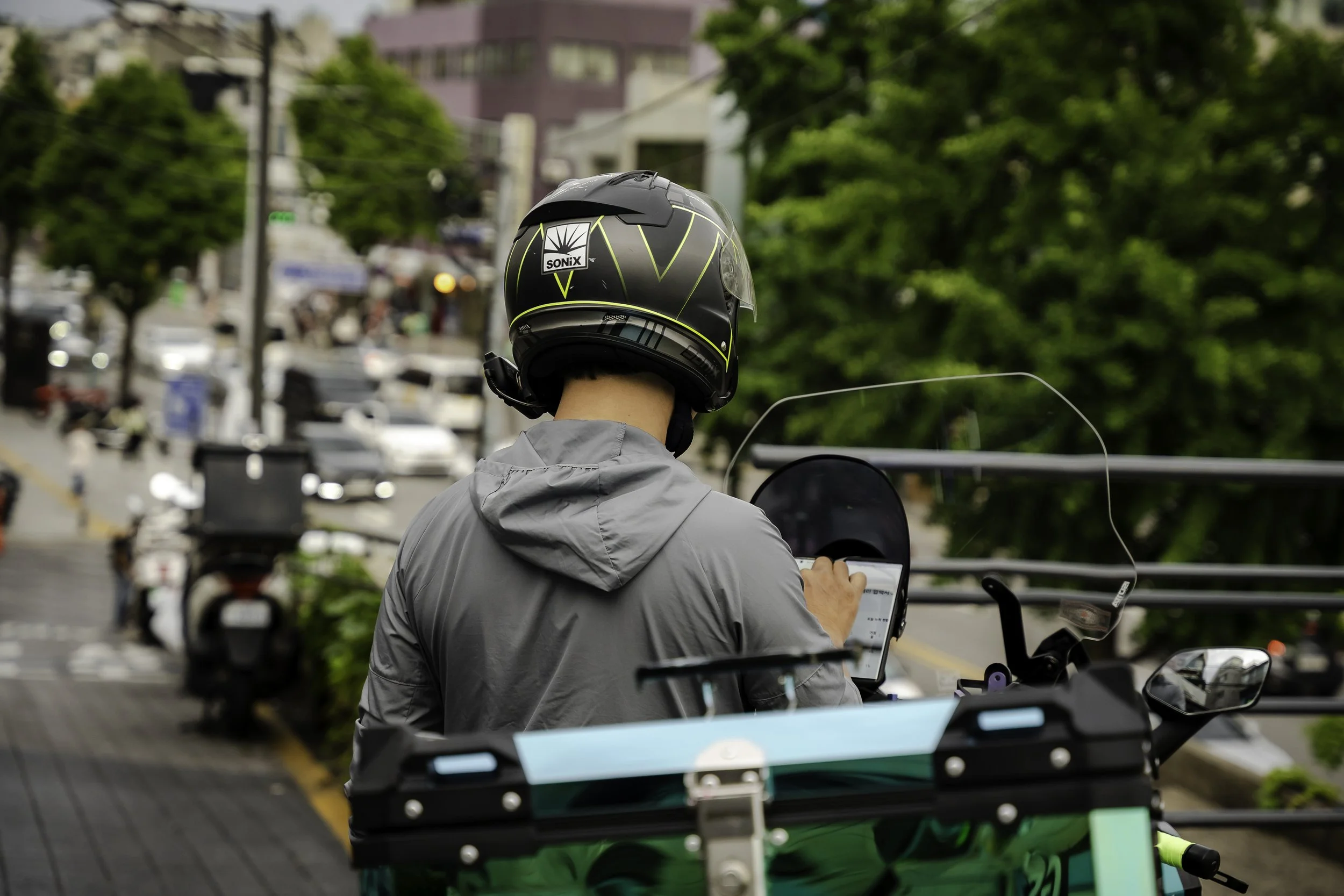 A person wearing a black helmet and a gray jacket, seen from behind, is using a smartphone while sitting on a green delivery bike on a city street, with cars and trees in the background.