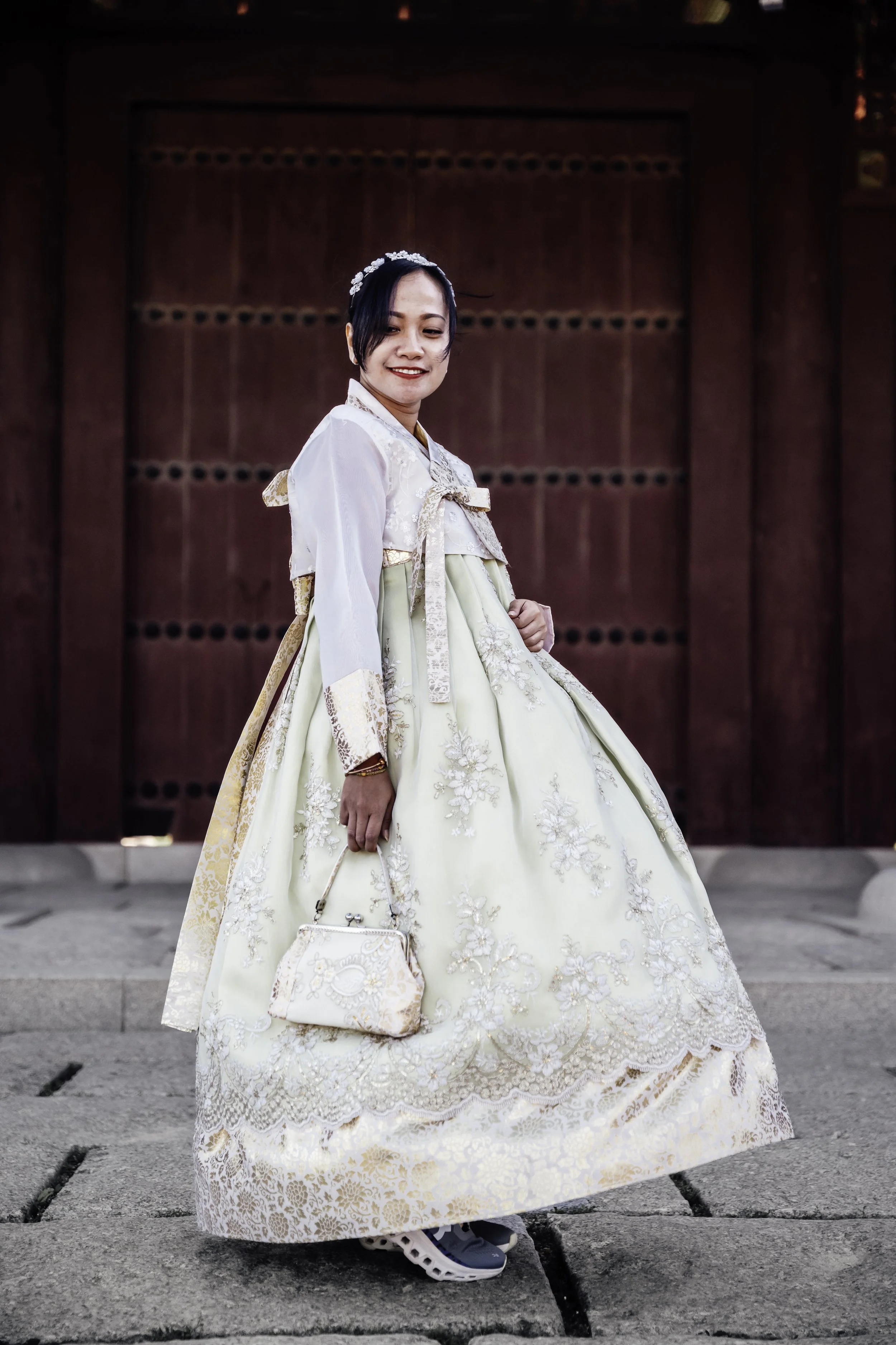 A woman wearing a traditional Korean hanbok, standing on stone pavement in front of a large wooden door.