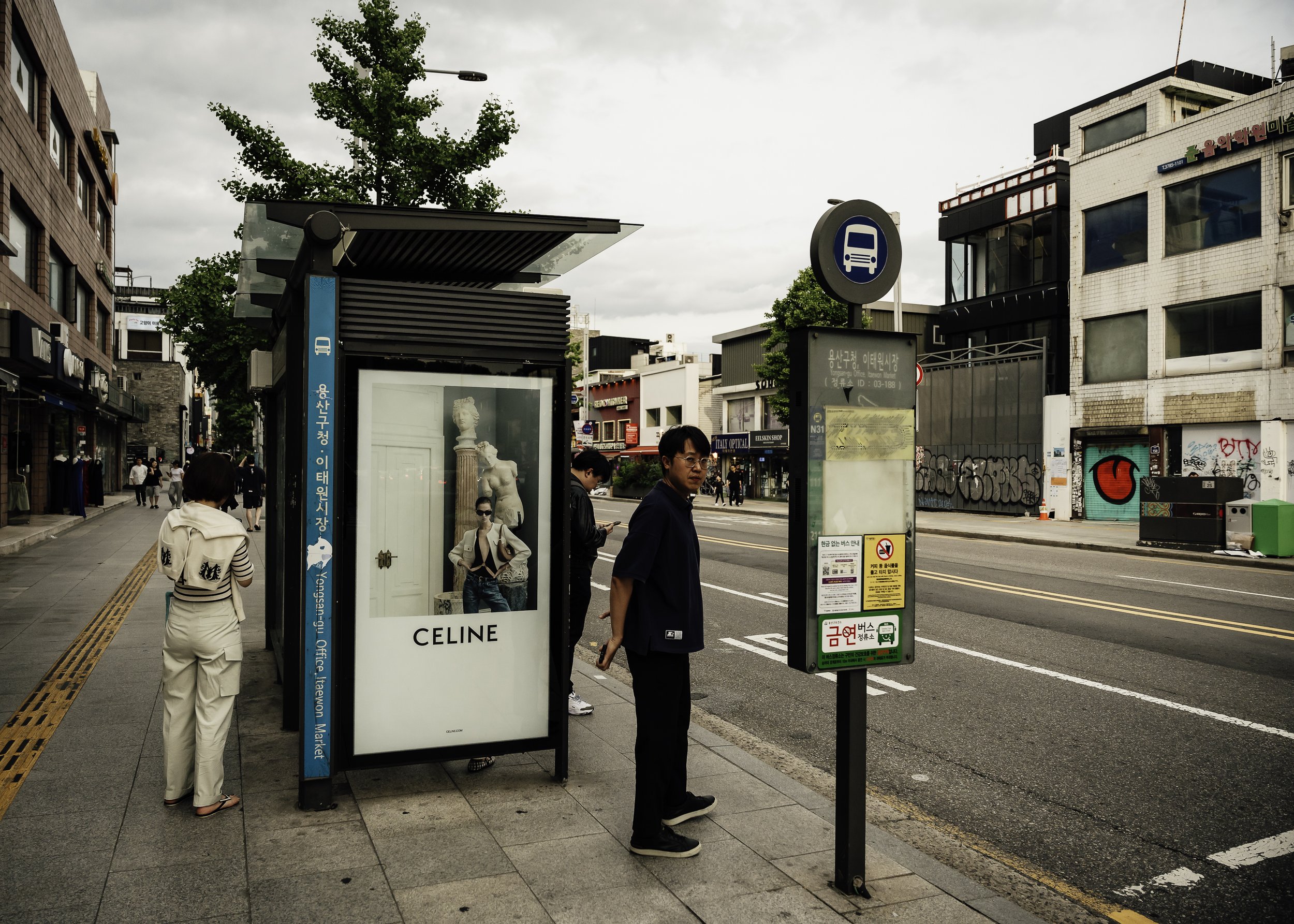People waiting at a bus stop on a city sidewalk with buildings and graffiti in the background.