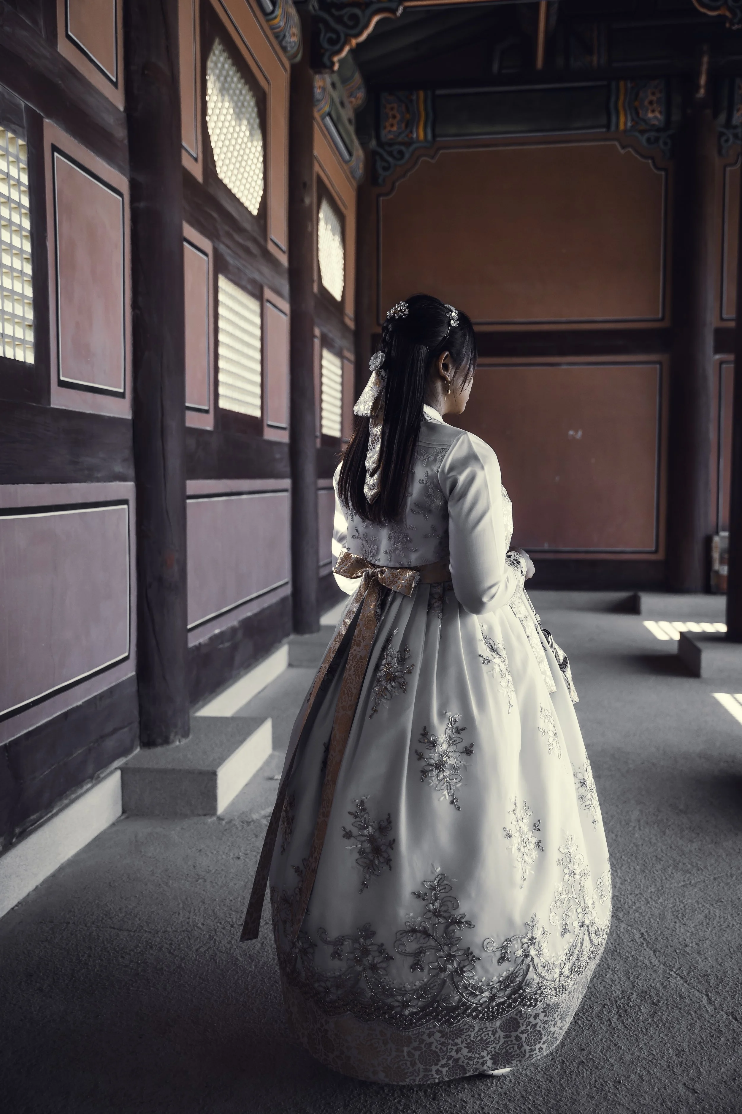 A girl wearing a traditional Korean hanbok stands inside a wooden building, facing away from the camera, with sunlight streaming through the windows.