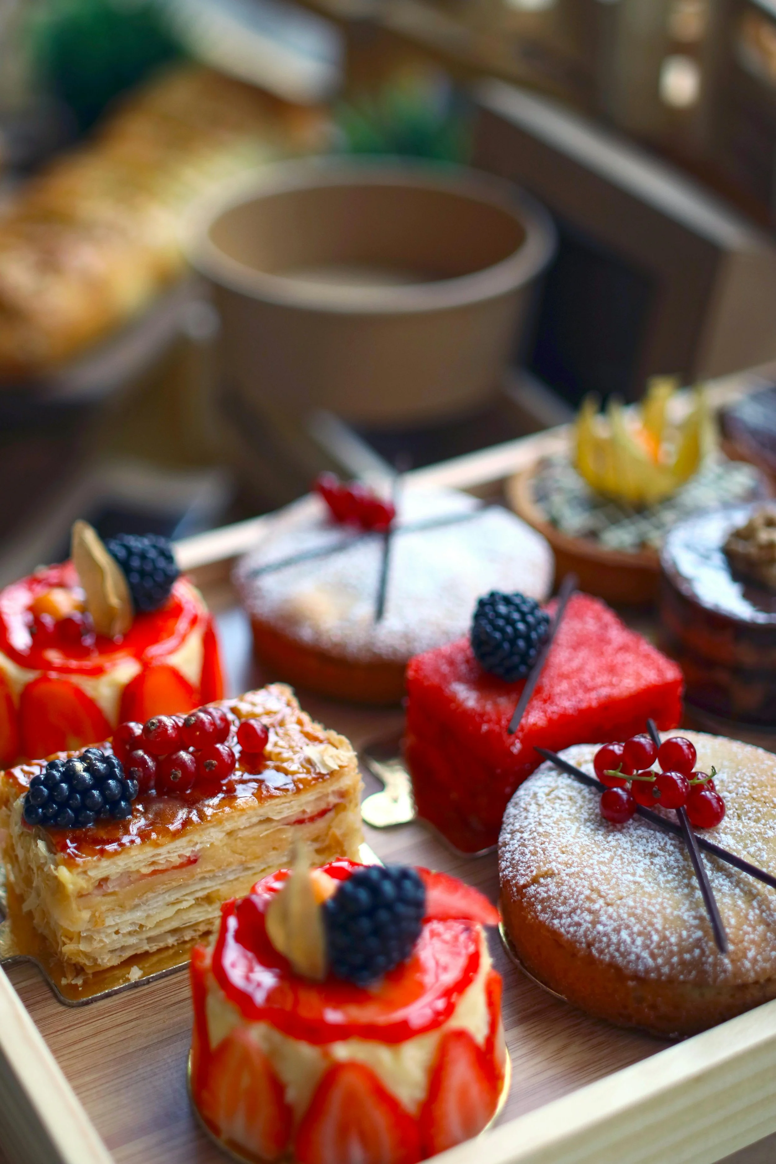 An assortment of colorful and elaborately decorated individual cakes and pastries topped with berries, fruit, and chocolate on a wooden tray.
