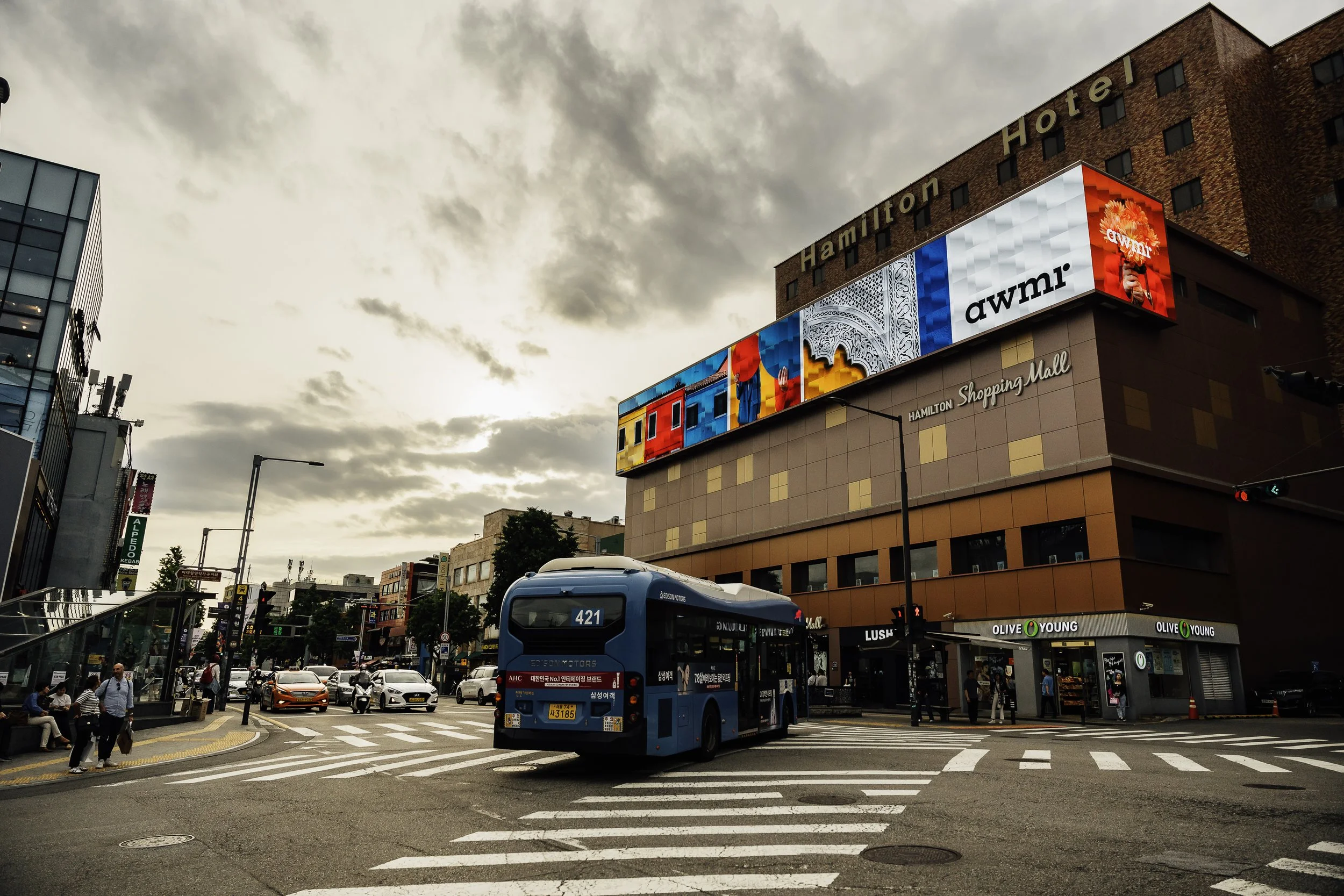 City crosswalk scene with pedestrians waiting, a blue bus crossing, and buildings including Hamilton Hotel in the background, with digital billboards on the building displaying colorful images and text.