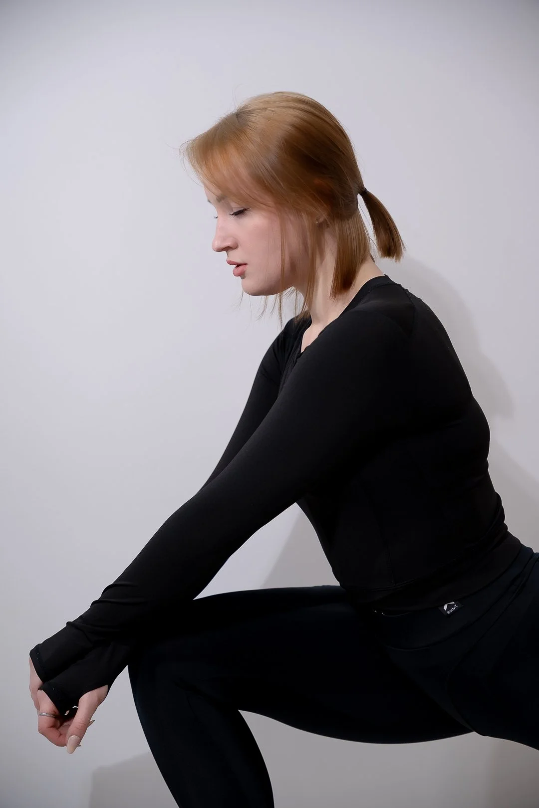 A woman with red hair tied in a small ponytail is stretching her leg while squatting, wearing a black athletic outfit against a plain white wall.