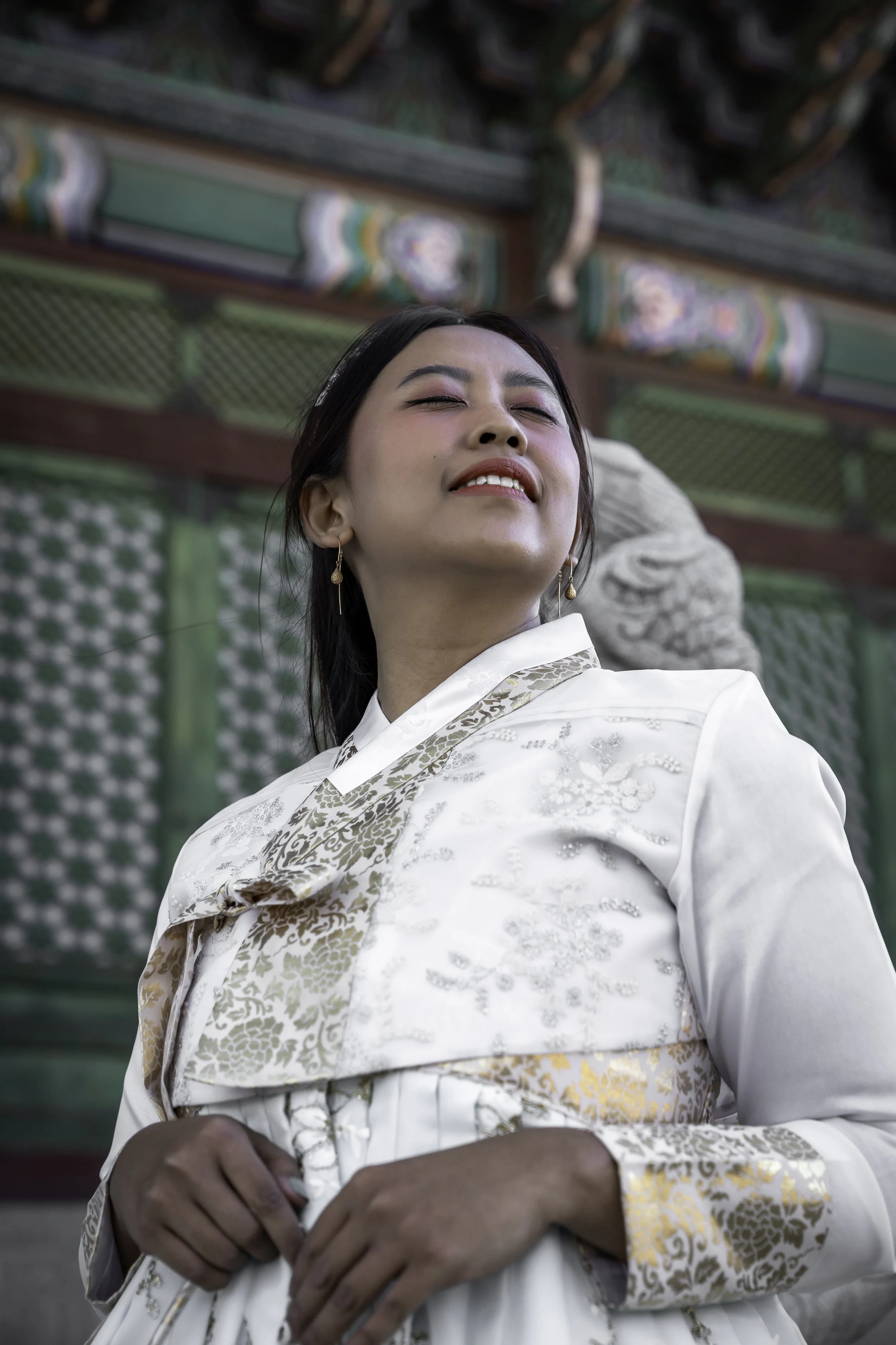 A woman in traditional attire smiling with her eyes closed, standing in front of colorful, ornate architecture.