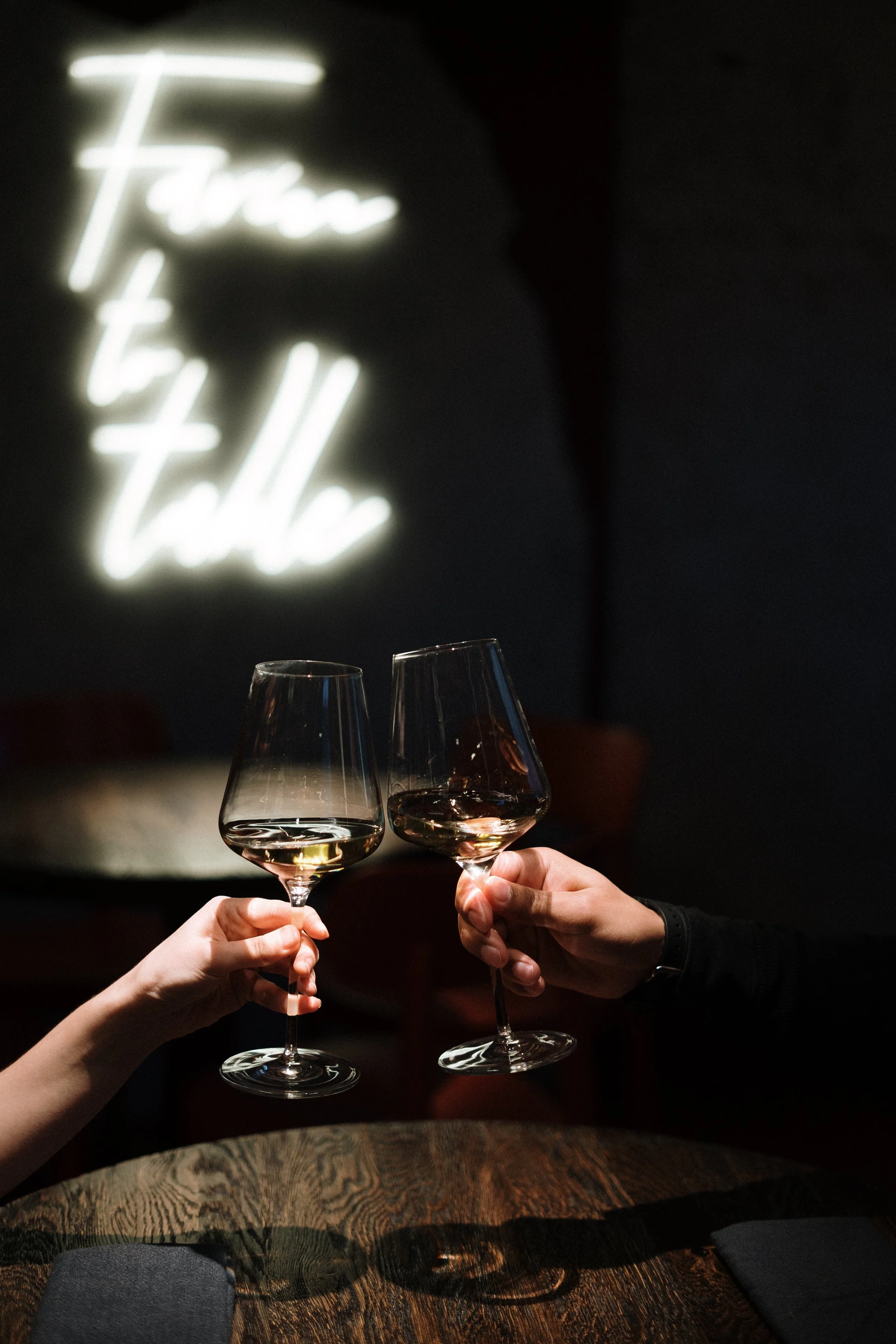 Two people clinking glasses of rosé wine at a dark, cozy table with a partially visible black napkin, in a setting with a blurred neon sign in the background that reads "Farn Talle."