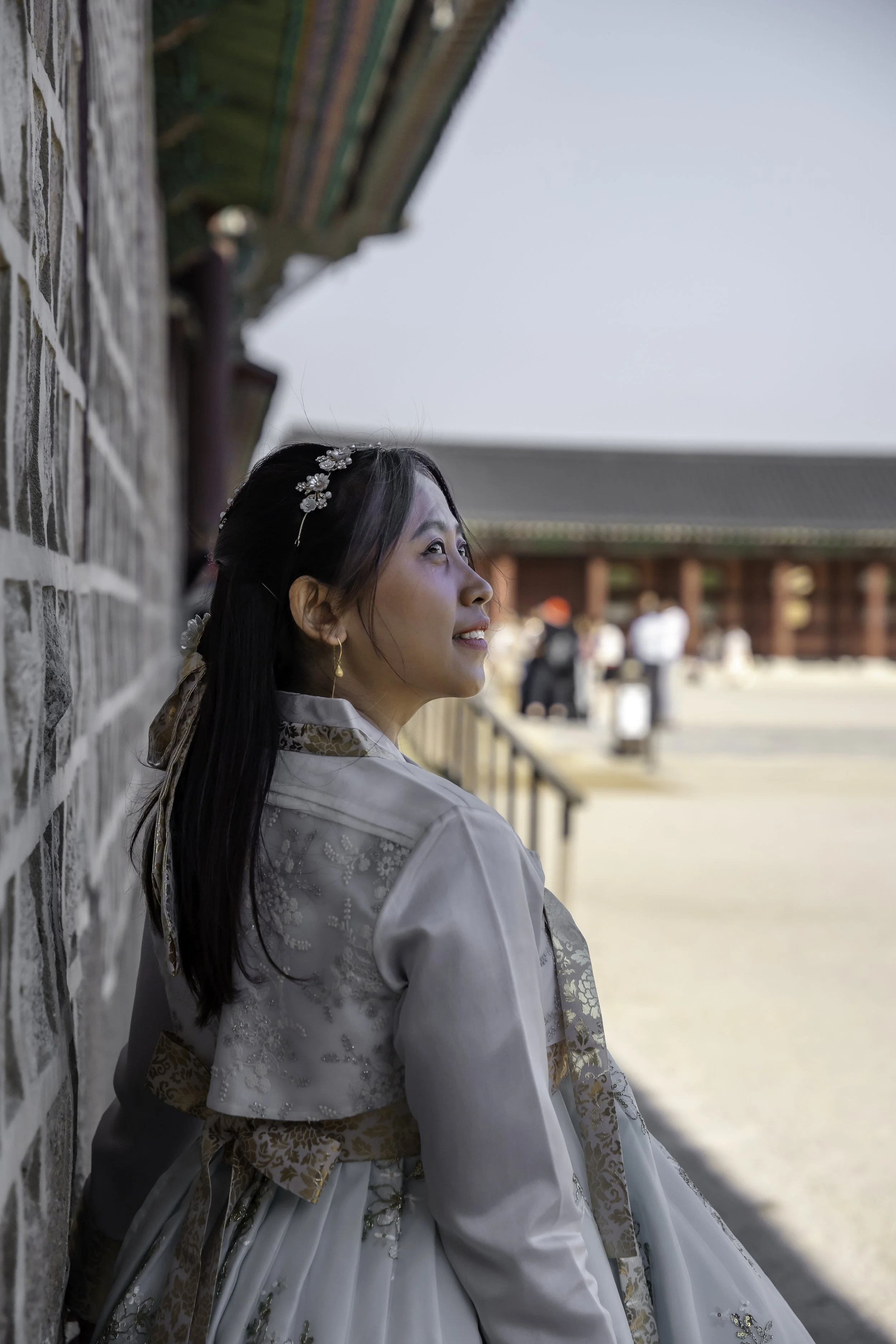 A woman with long dark hair, wearing a traditional Korean hanbok, leaning against a stone wall outdoors, looking upward with a serene expression. In the background, a traditional Korean building and a few people are visible.