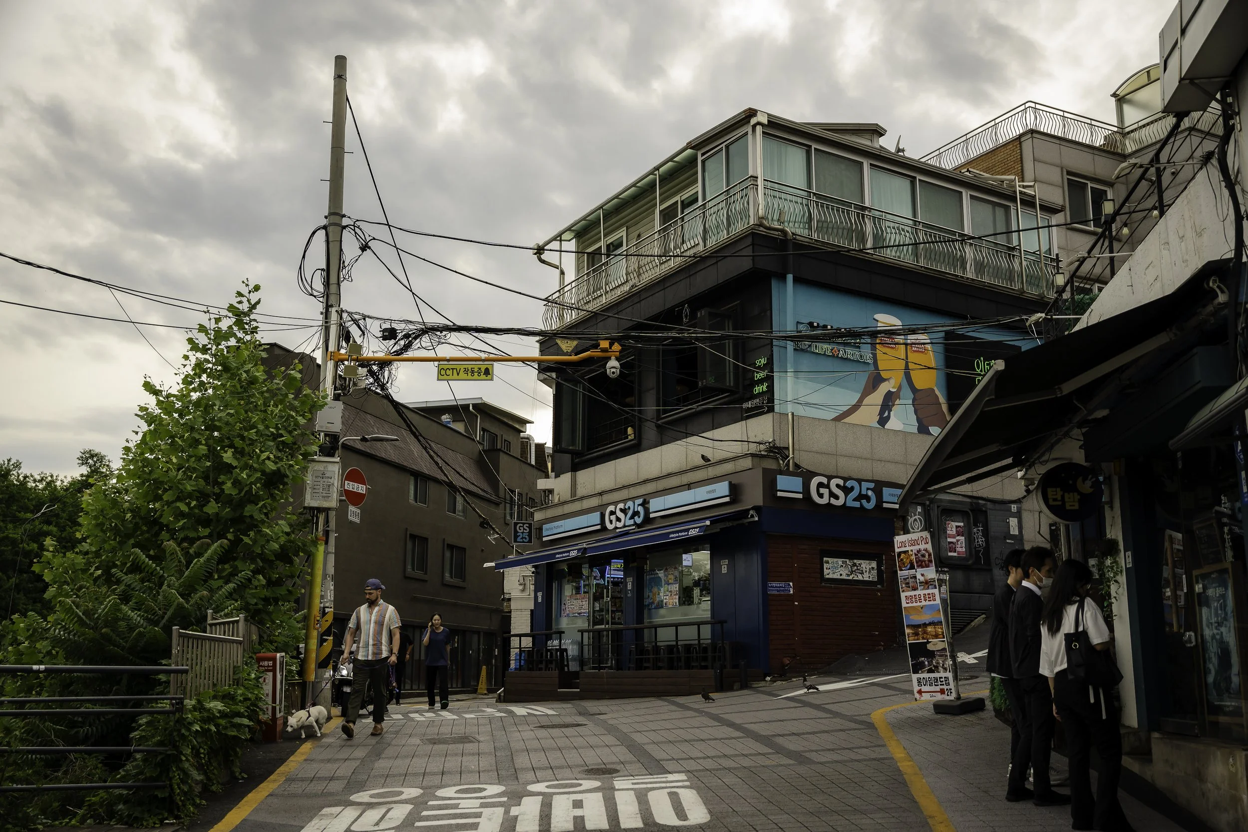 Street scene with GS25 convenience store, pedestrians, power lines, and a building with a mural on a cloudy day.