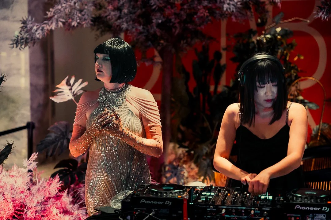 Two women, one dressed in a sparkling gown with gloves and pearls, and the other in a black dress with headphones, are behind a DJ console at a stylish event with pink and red floral decorations.