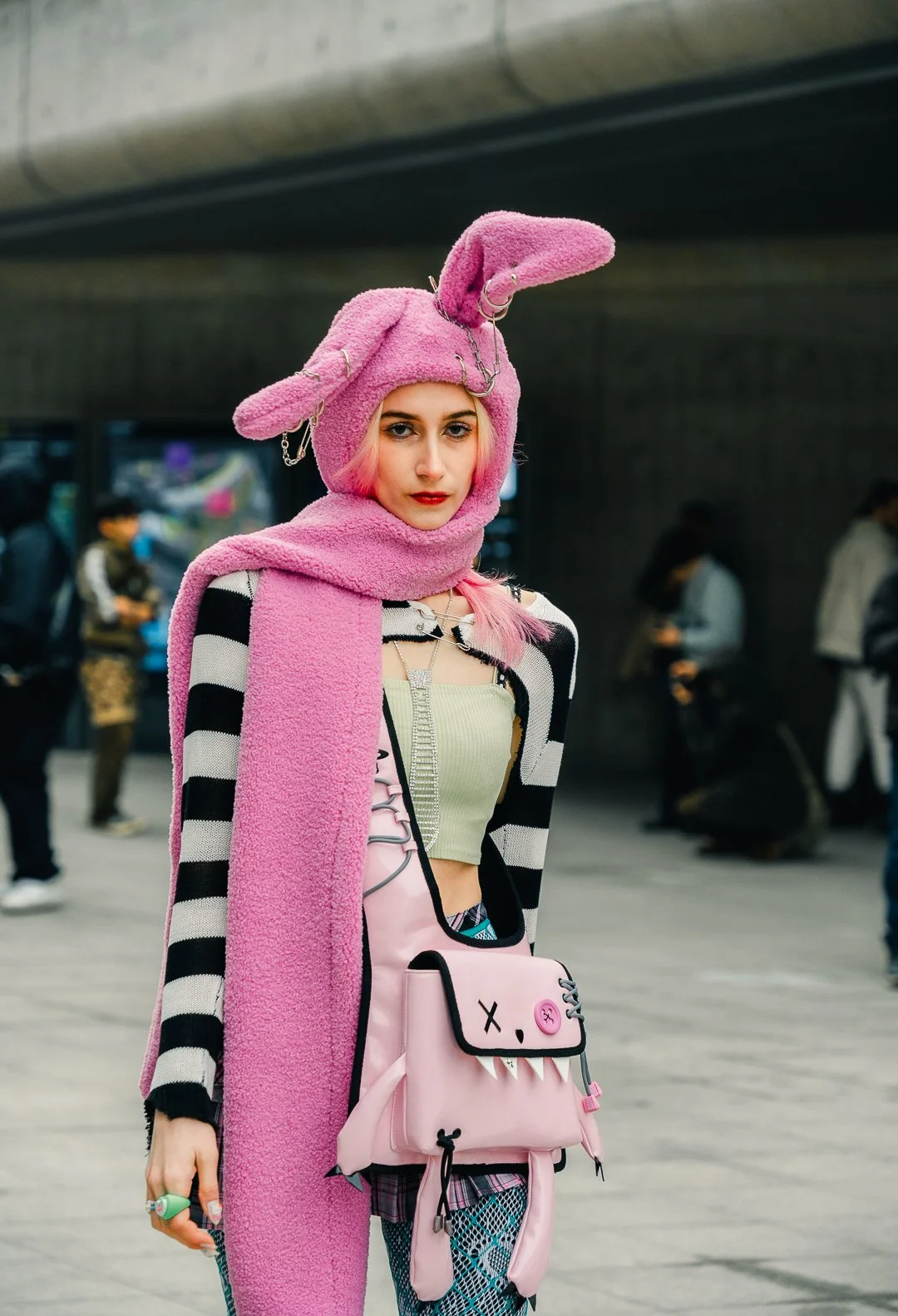 Young woman with pink hair wearing a pink bunny hat and scarf, striped black and white jacket, and carrying a pink bag with a monster face design, in an urban setting.