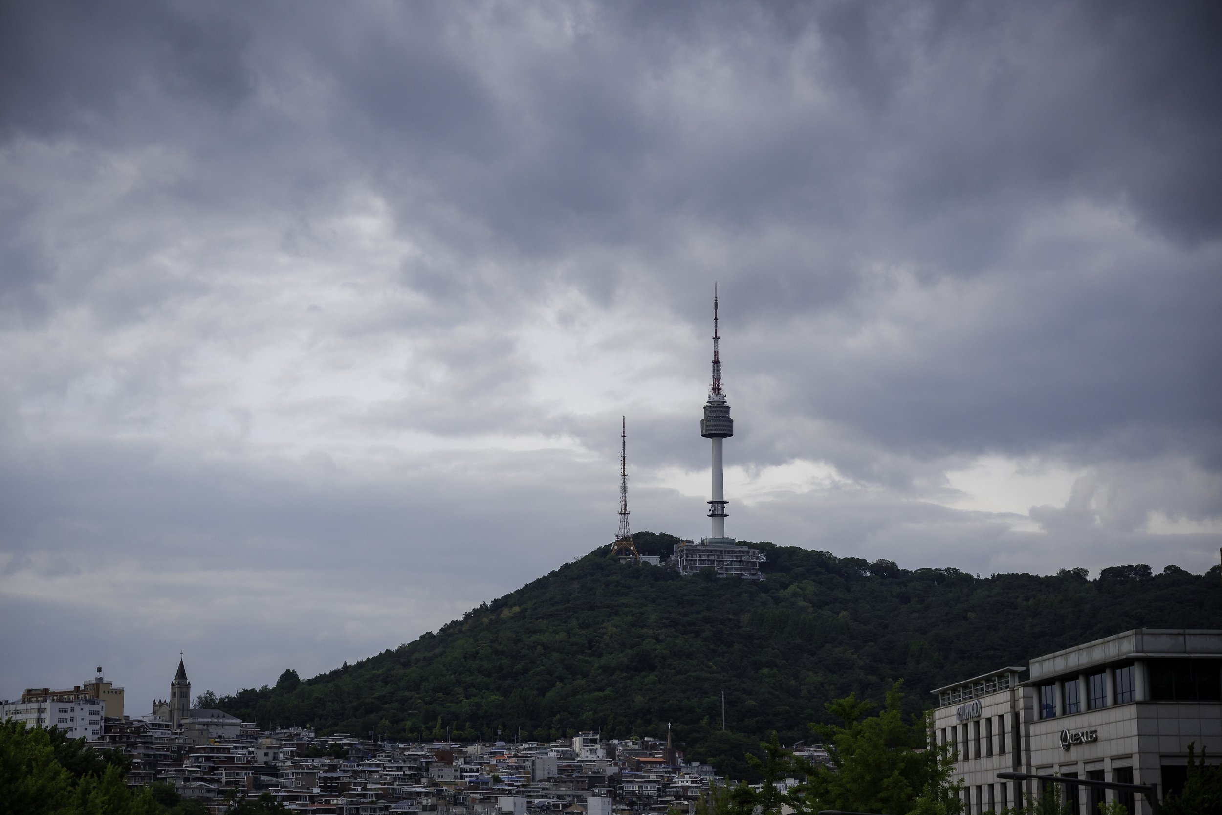 View of a city with a hill topped by a tall communication tower and antennas, under a cloudy sky.